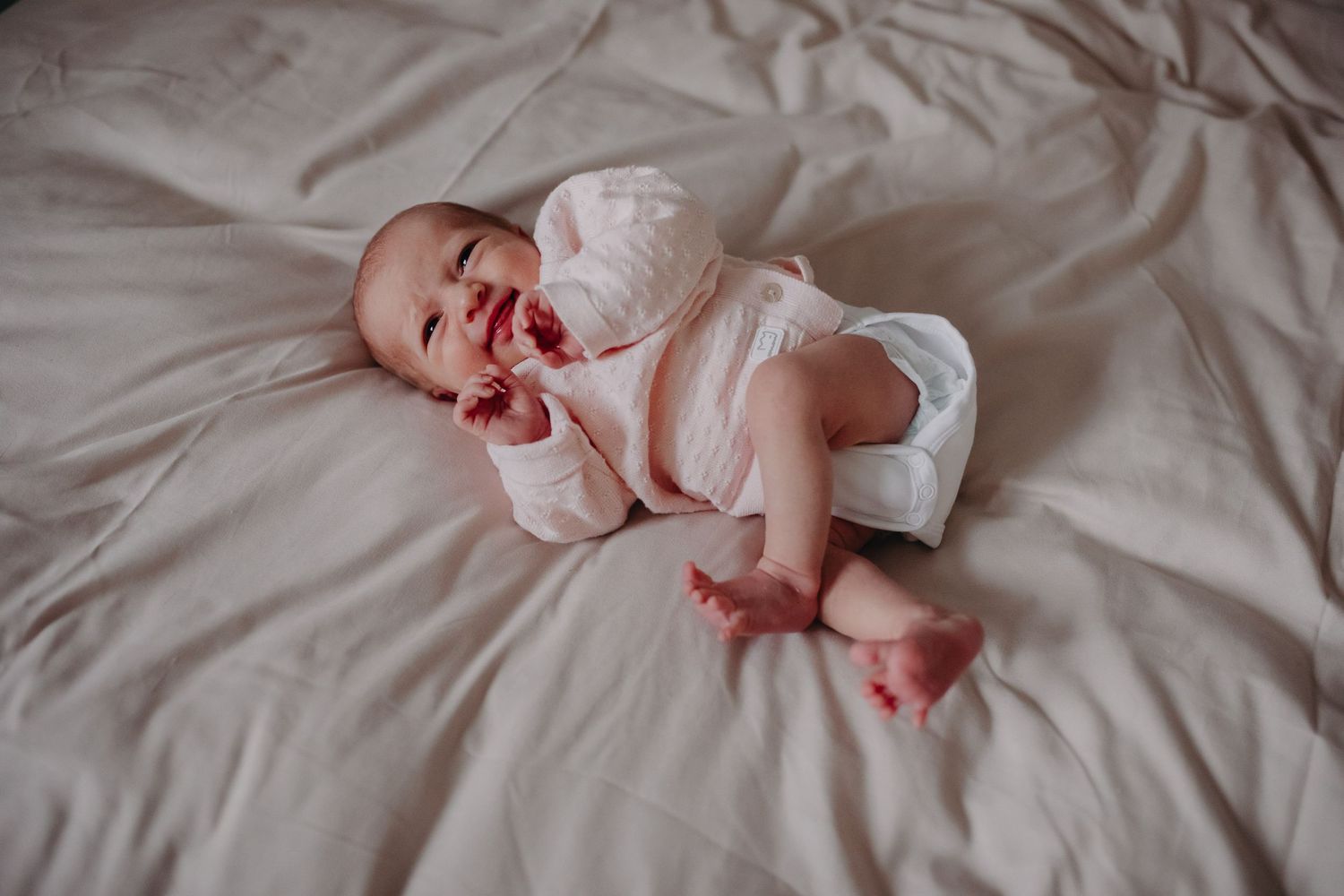 A newborn baby wearing white clothing yawns while lying on light gray bedding.