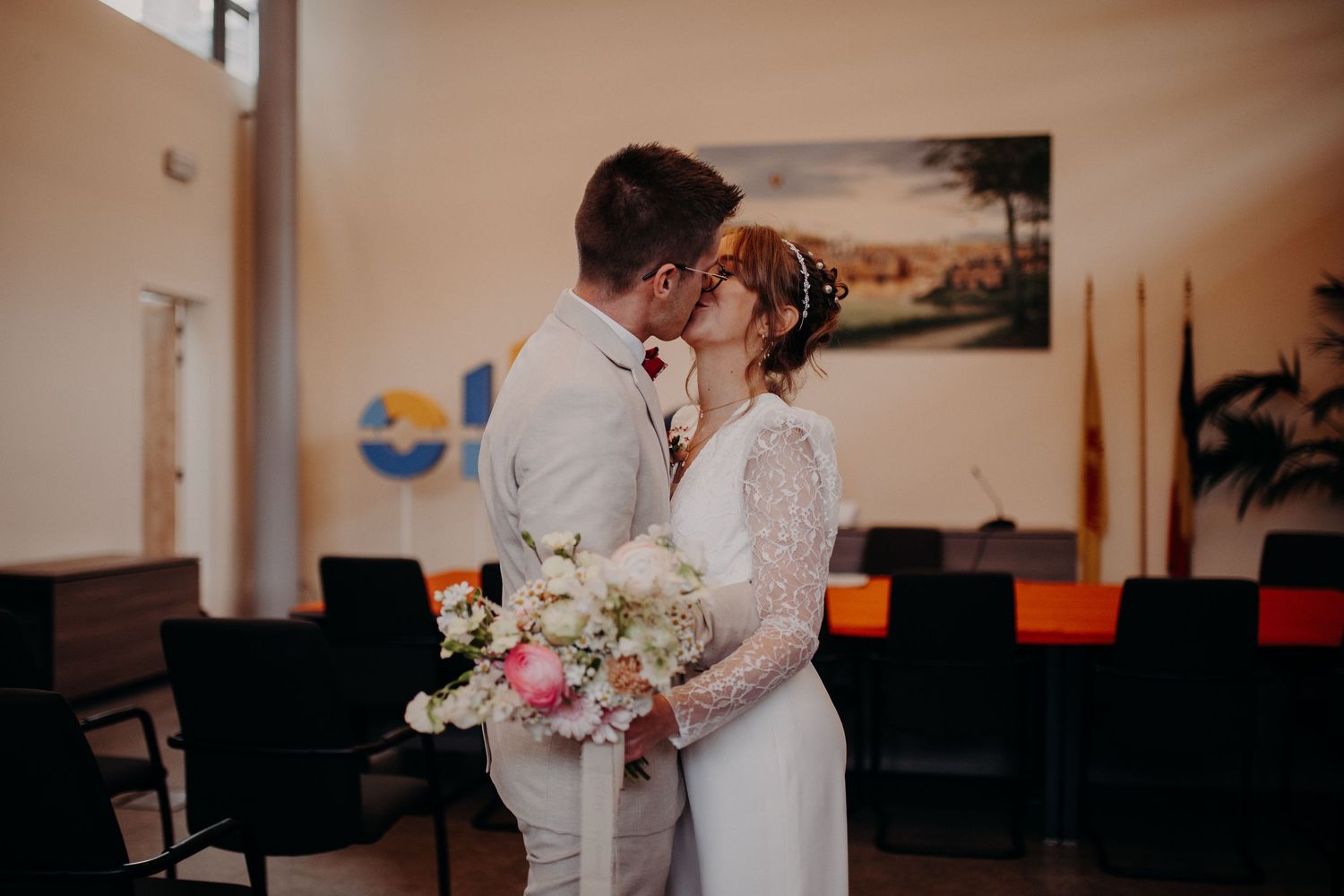 A romantic wedding scene with a couple sharing a kiss while holding a pink and white floral bouquet in an indoor setting.