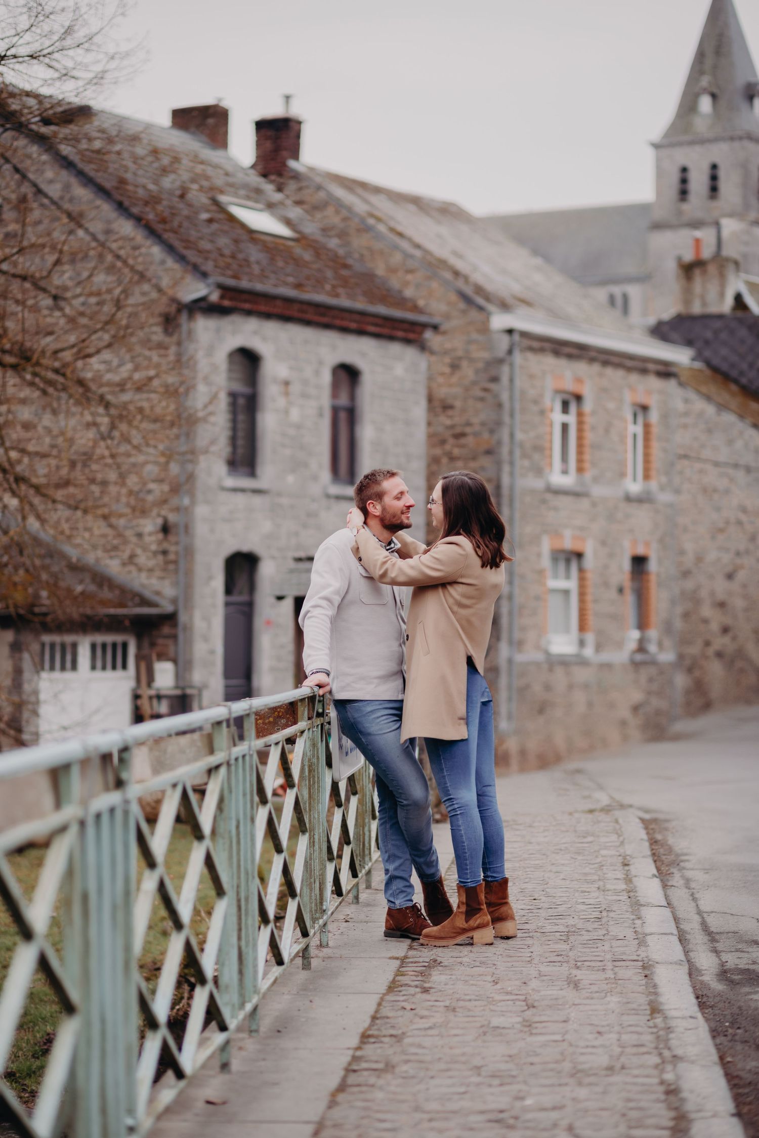 A romantic couple stands together on a cobblestone bridge in a quaint European village with stone buildings and church spire.