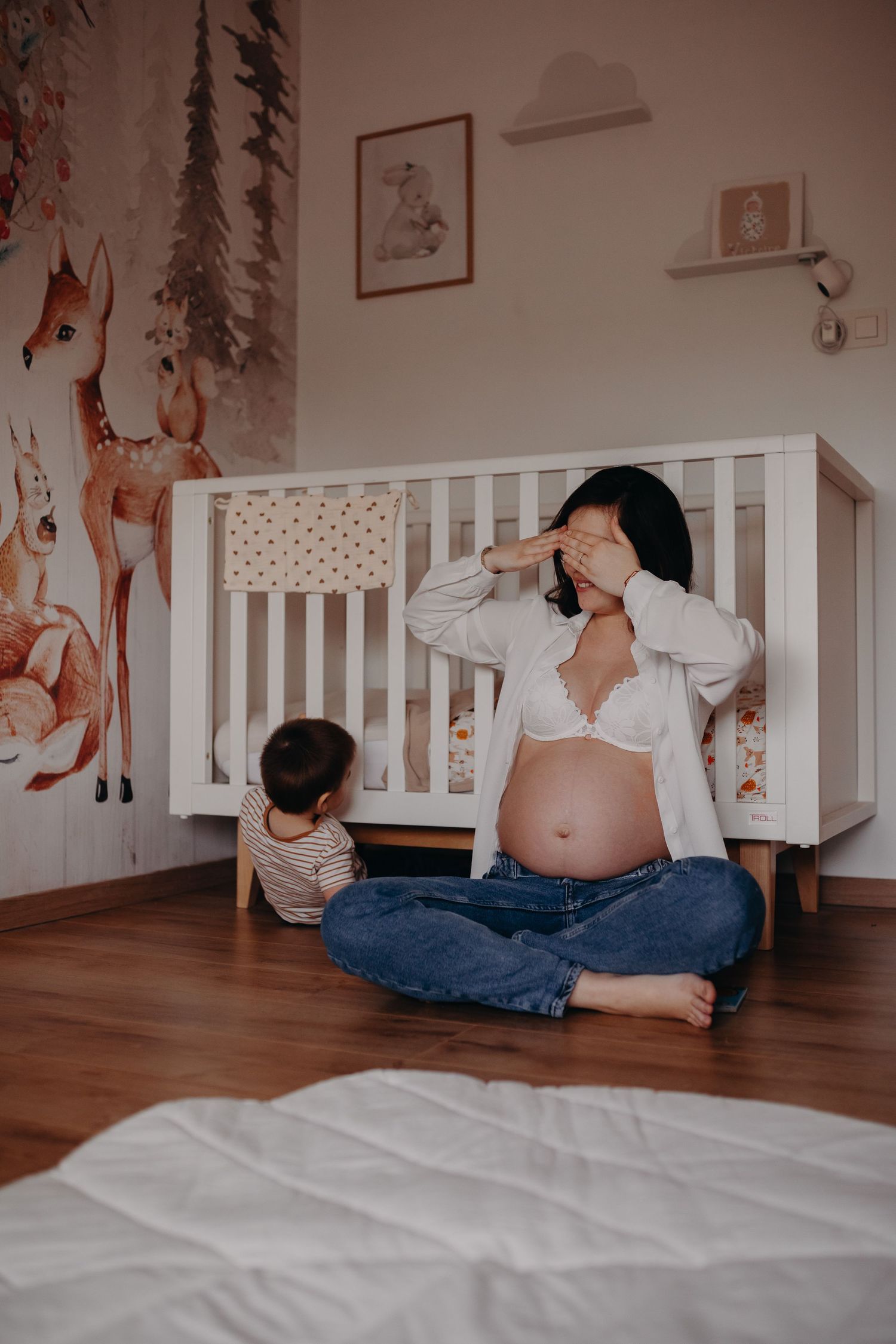 A nursery with woodland mural wallpaper features a white crib and person sitting with toddler on hardwood floor.
