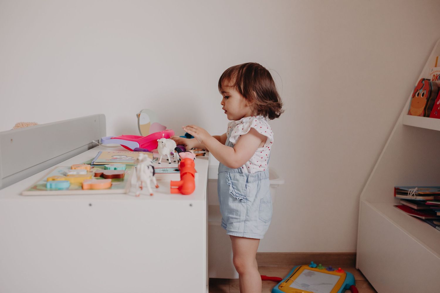 A child in denim overalls plays at a white table with colorful toys and art supplies.