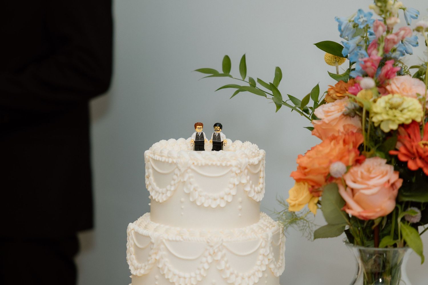 Tiered wedding cake with two grooms cake toppers beside a floral arrangement.