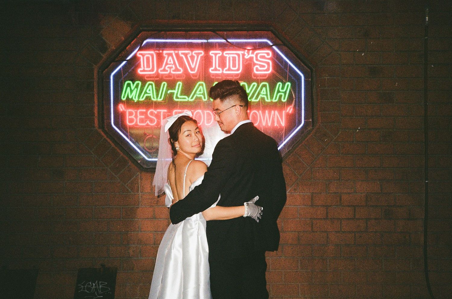 Bride and groom embracing beneath a colorful vintage neon sign at a brick-walled retro venue, captured on film.