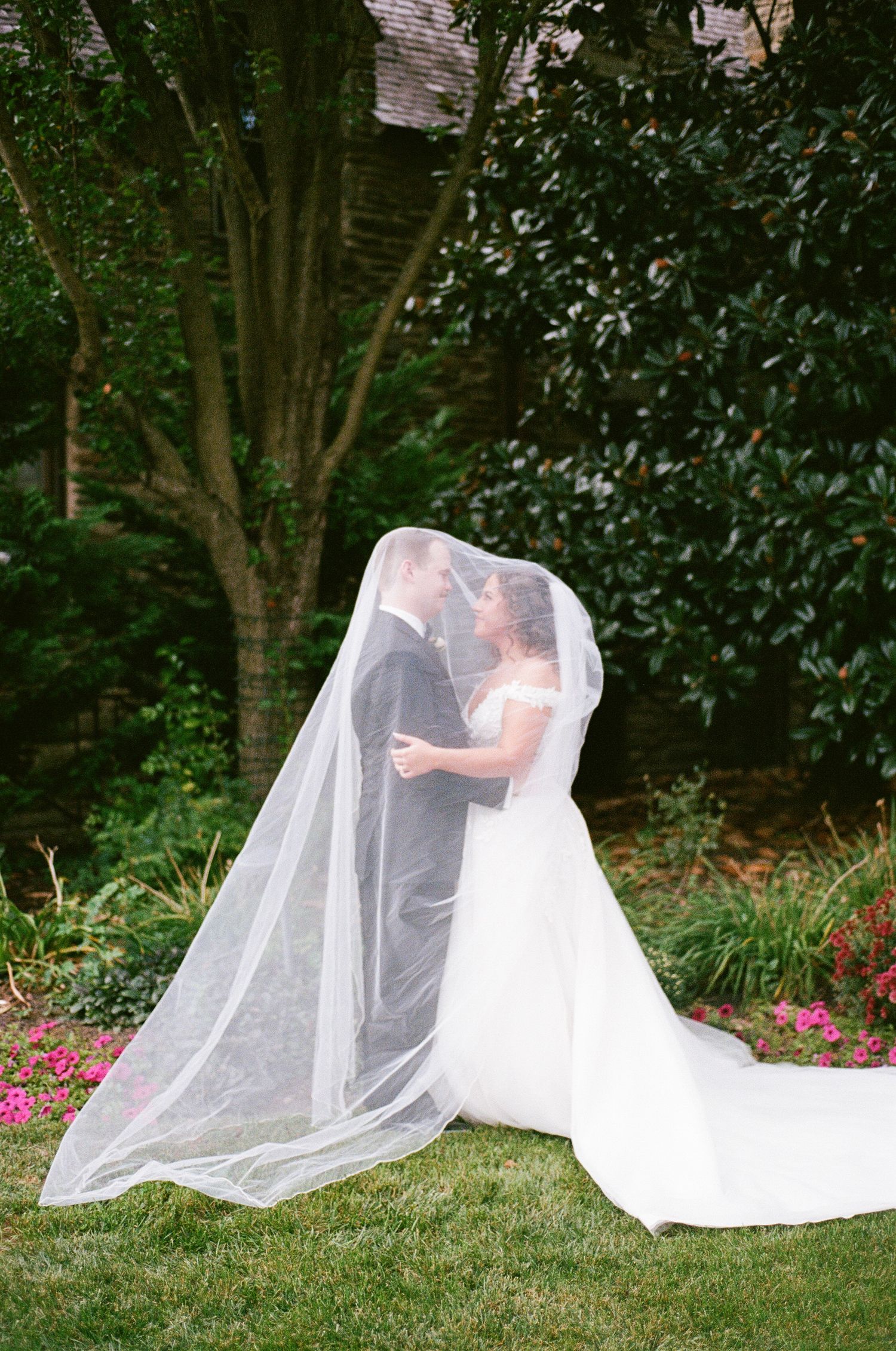 Bride photographed from behind showing the full length of her veil and gown in a lush garden.