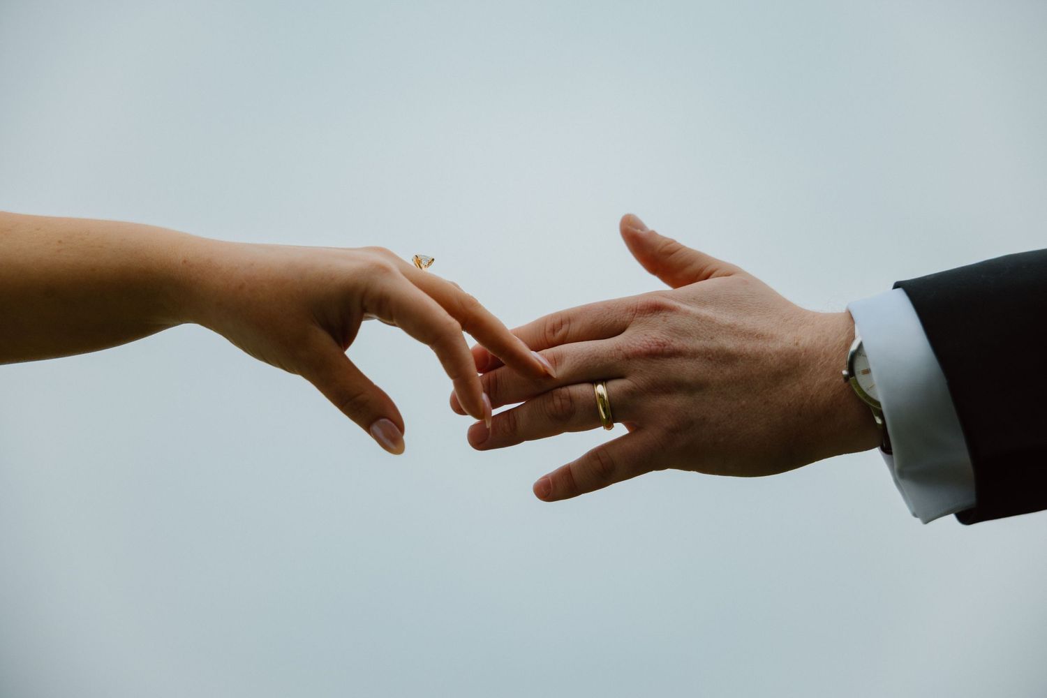 Bride and groom hands reaching toward each other showing wedding ring against soft blue backdrop.