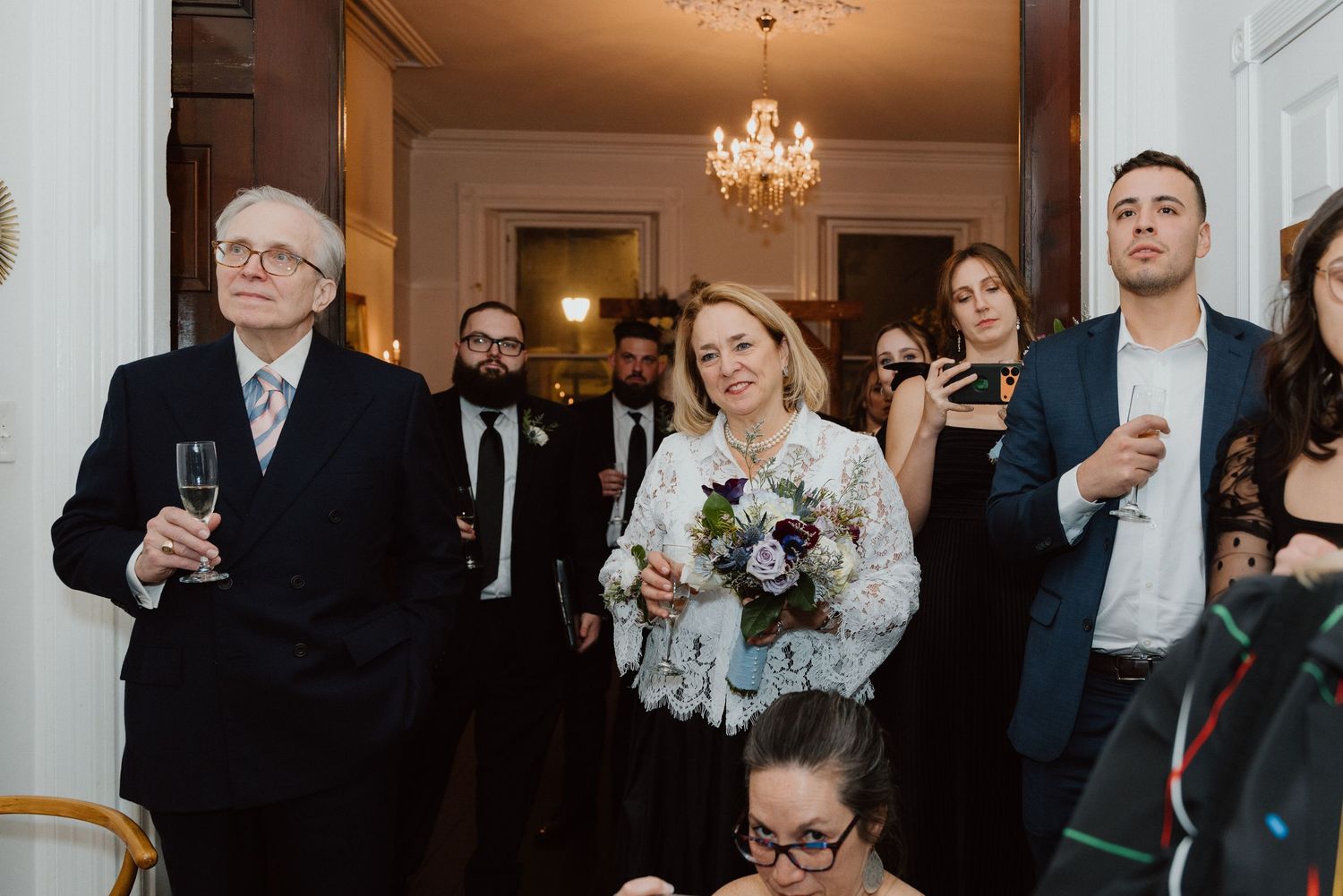 Guests raising champagne glasses during a heartfelt wedding toast beneath a chandelier in a historic venue.