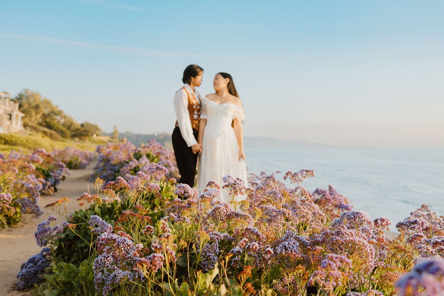 Two brides embracing on a coastal hillside blanketed with purple wildflowers at golden hour.