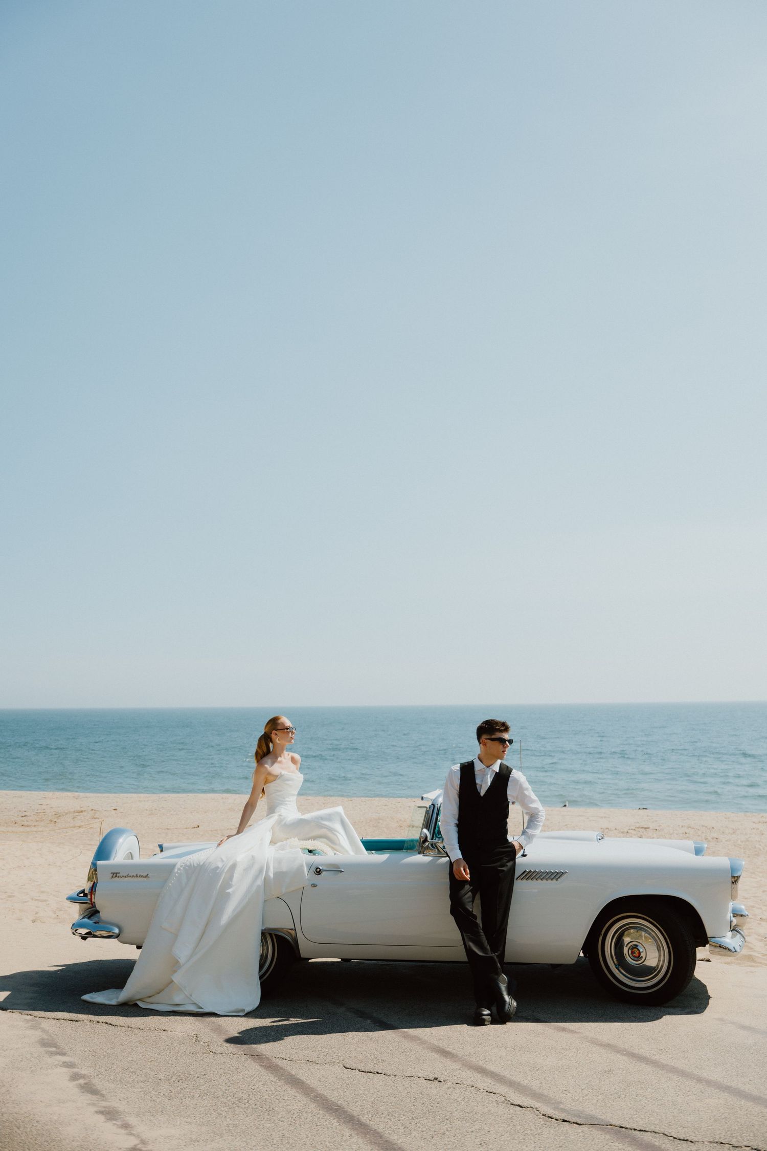 Bride and groom standing beside a vintage convertible along the oceanfront at sunset.