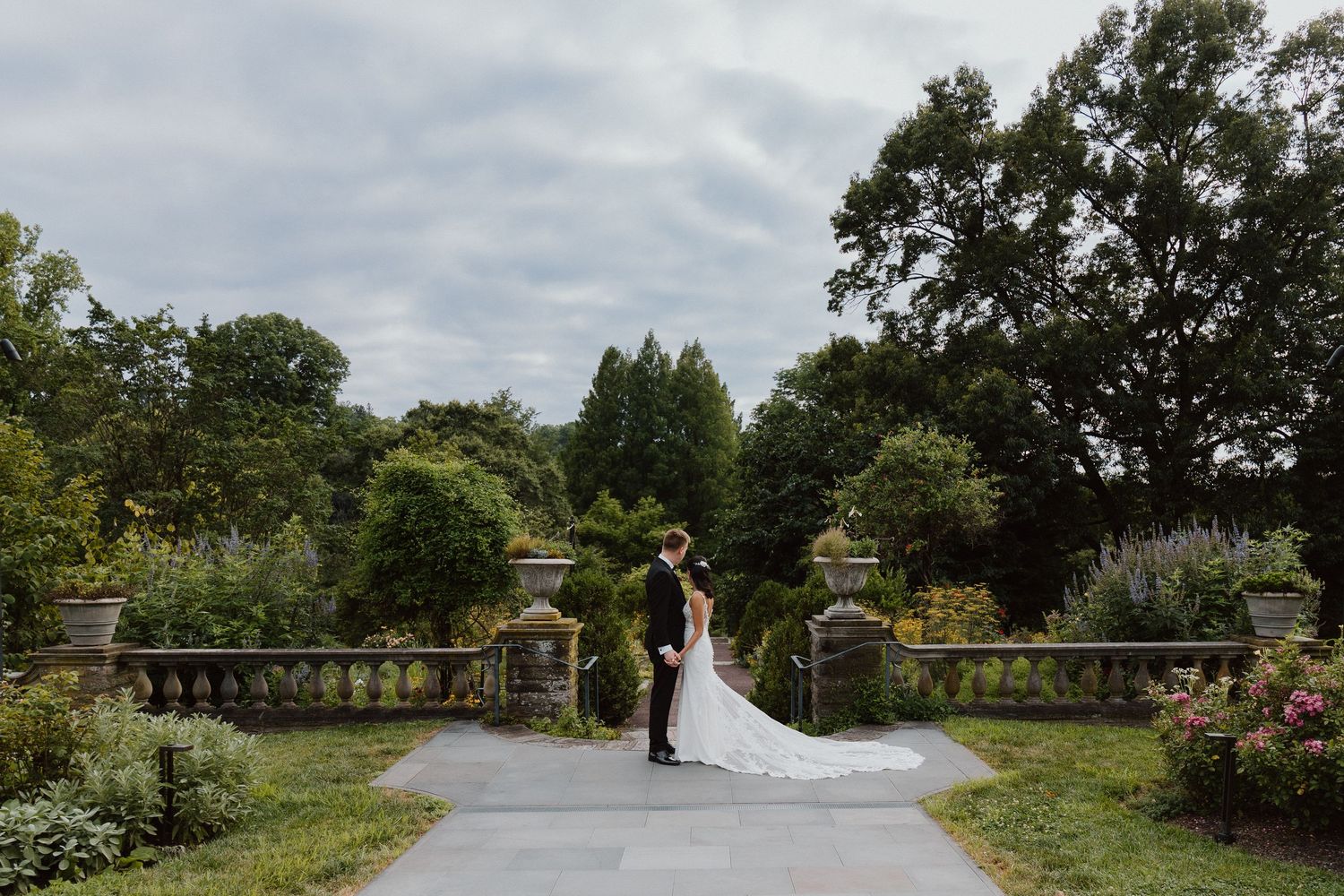 Bride in a flowing white gown standing on a stone garden path framed by arched garden gateways.