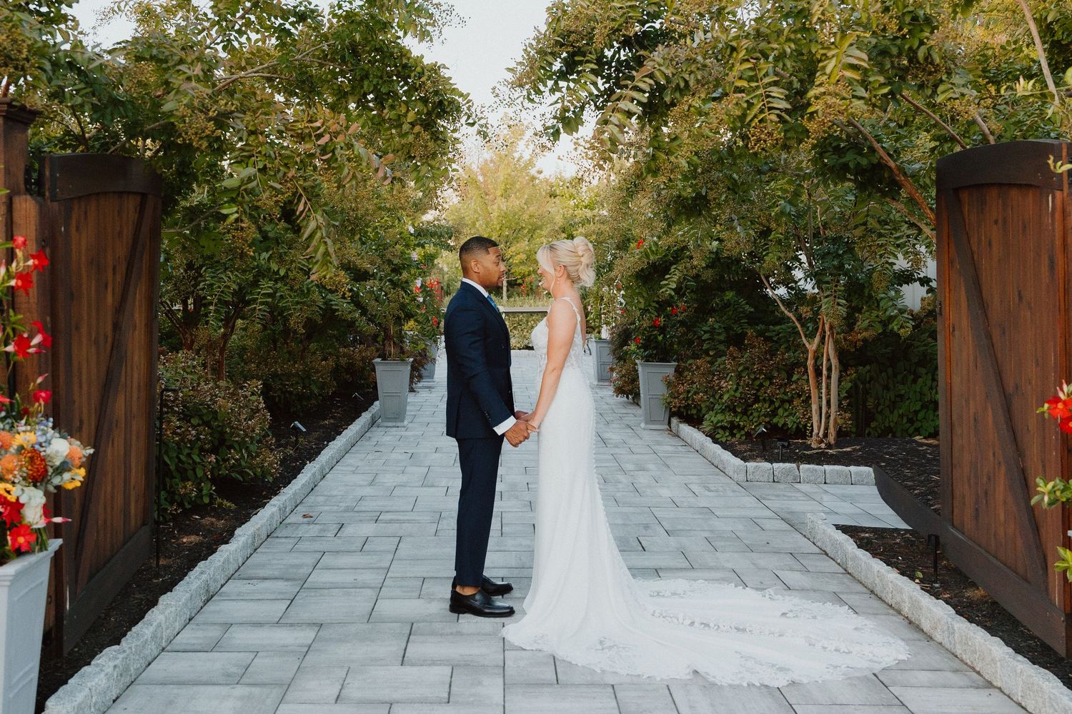 Bride and groom holding hands on a stone garden pathway framed by lush foliage.