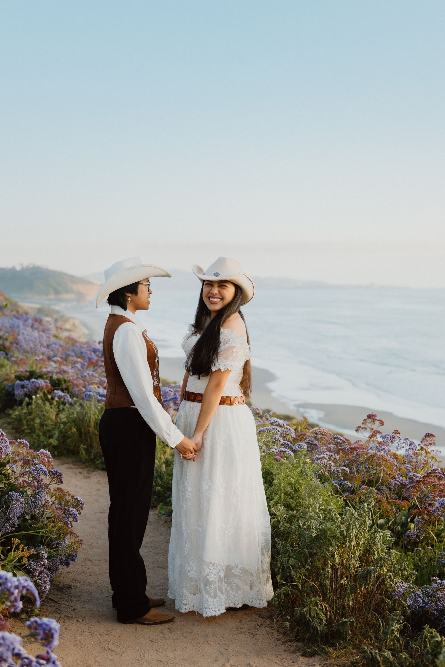 Couple holding hands on a coastal bluff with wildflowers during a seaside elopement.