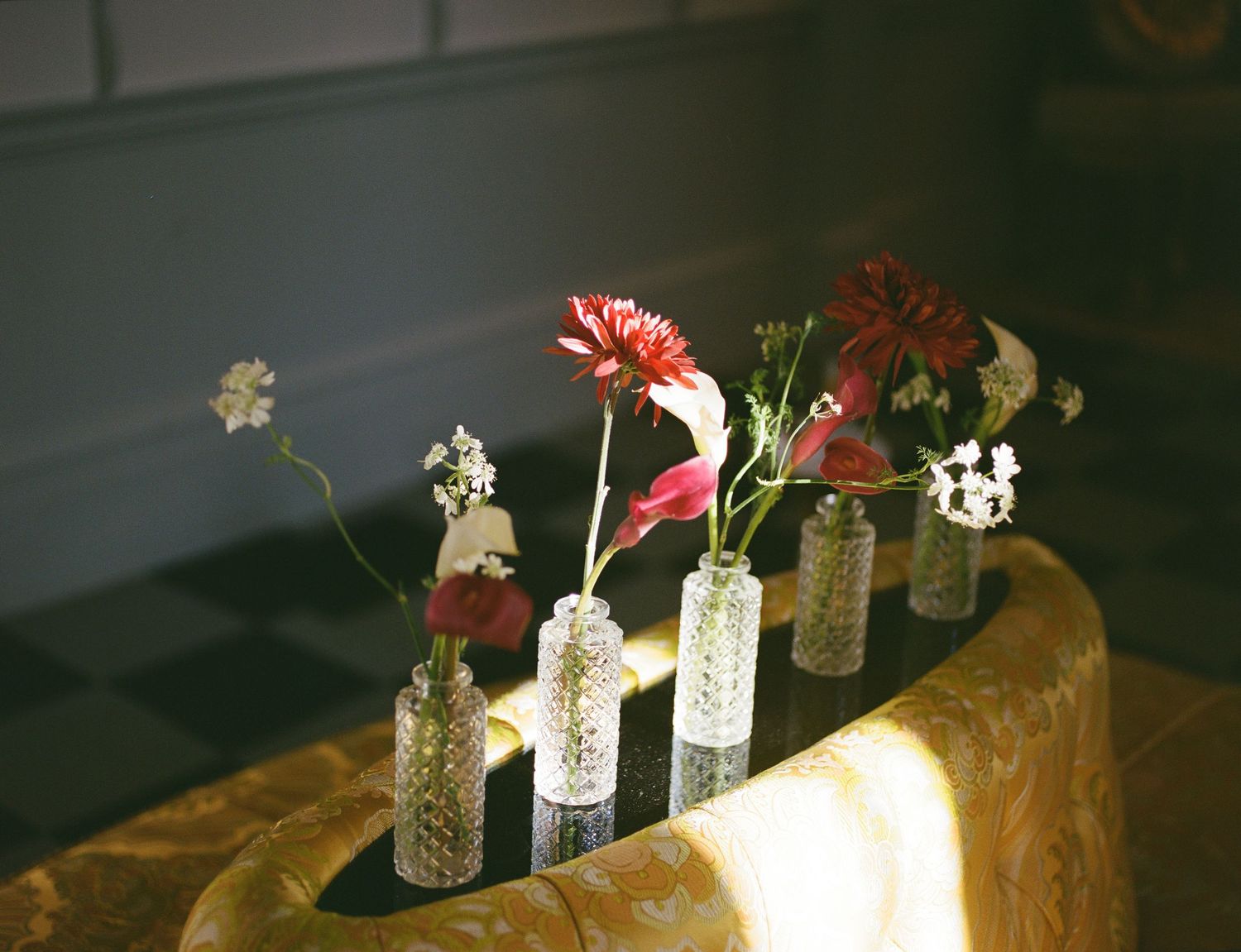 Reception table styled with assorted bud vases of red and yellow blooms beside candles and a glowing lamp.