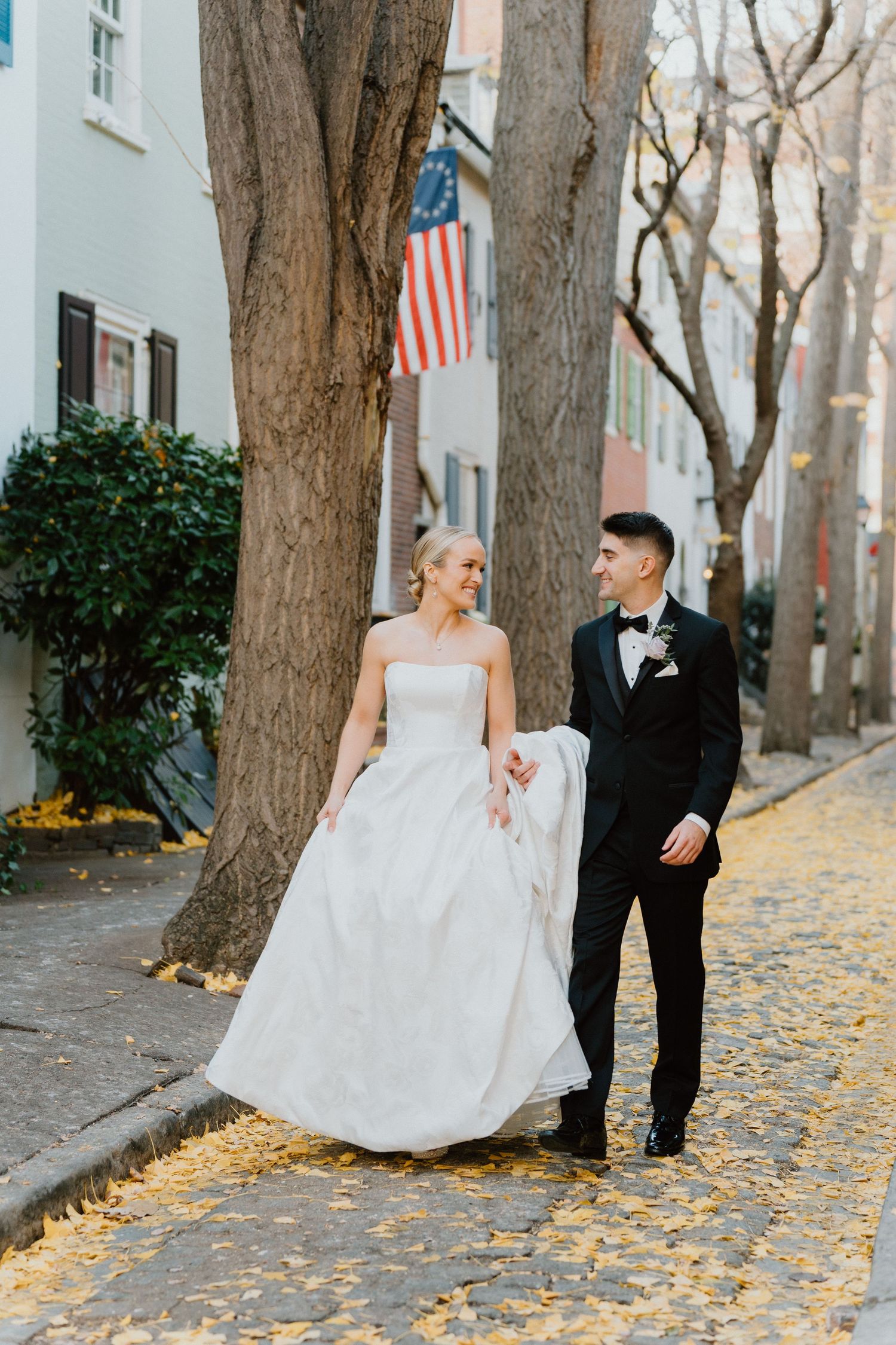 Bride and groom standing on a brick sidewalk in front of historic row homes with an American flag overhead.