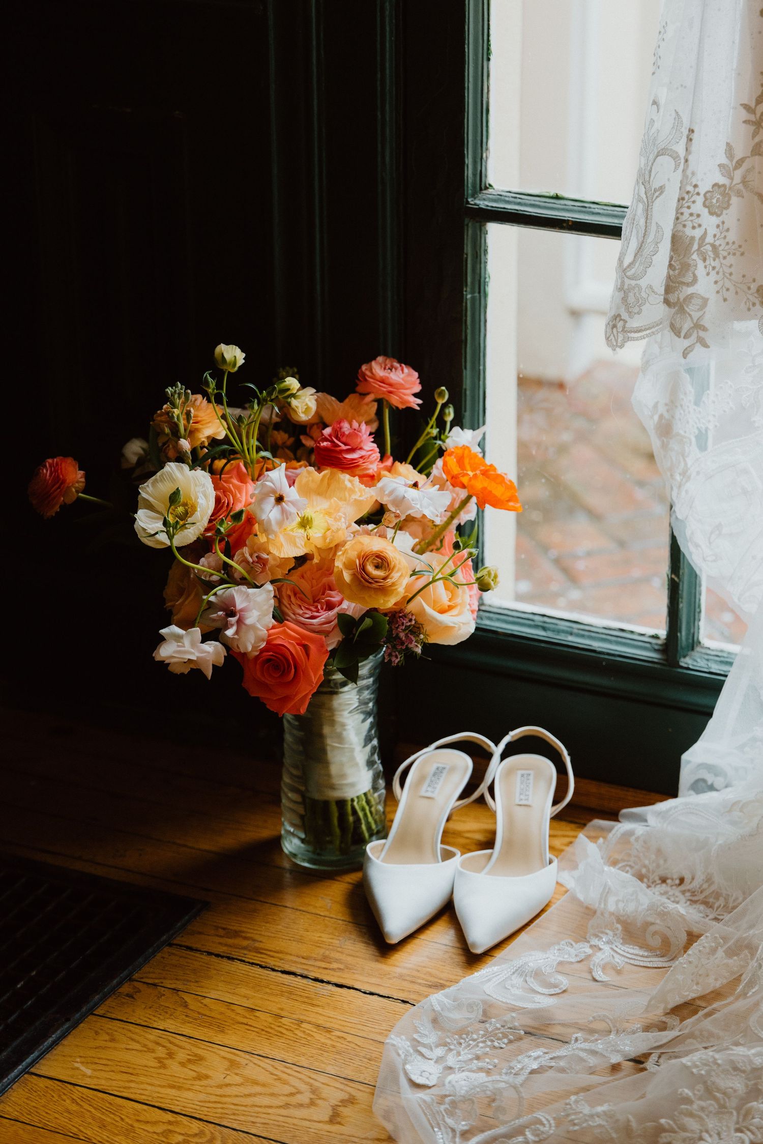 Bridal bouquet of coral ranunculus and white roses beside ivory wedding heels on hardwood floor.