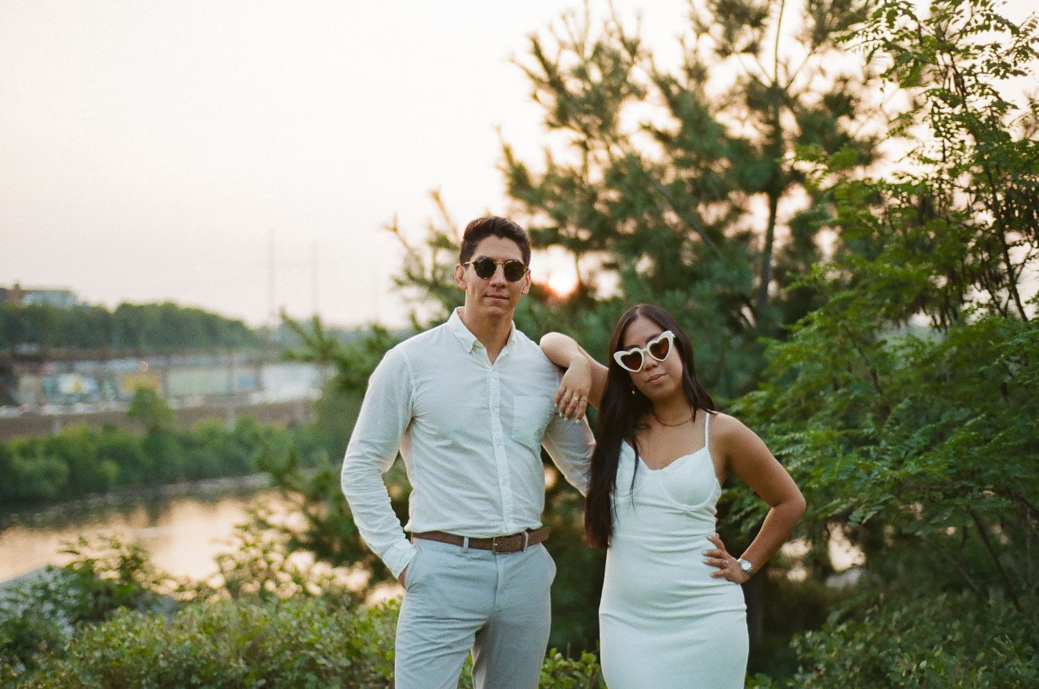 Couple in light outfits posing among trees on a sunny day.