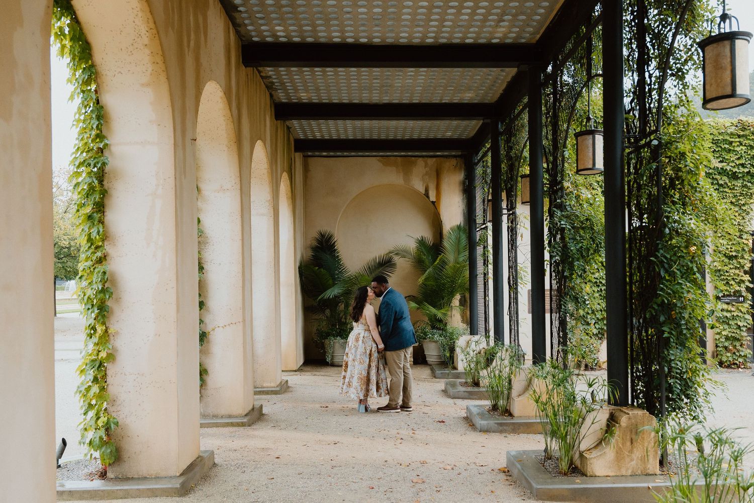 Bride and groom walking down a covered colonnade lined with arches and trailing greenery.
