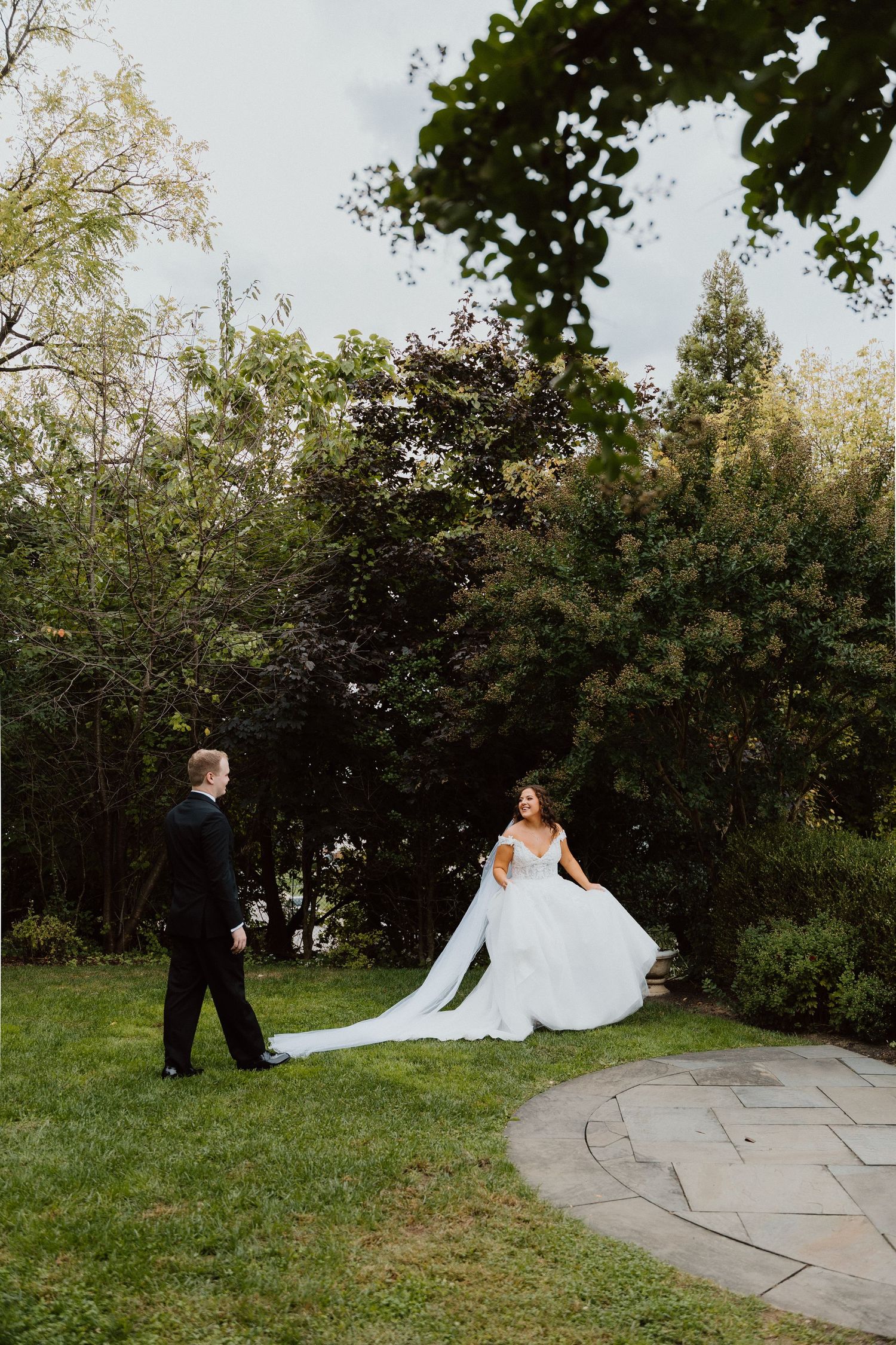 Bride in a long veil walking through a tree-lined garden path on the wedding day.