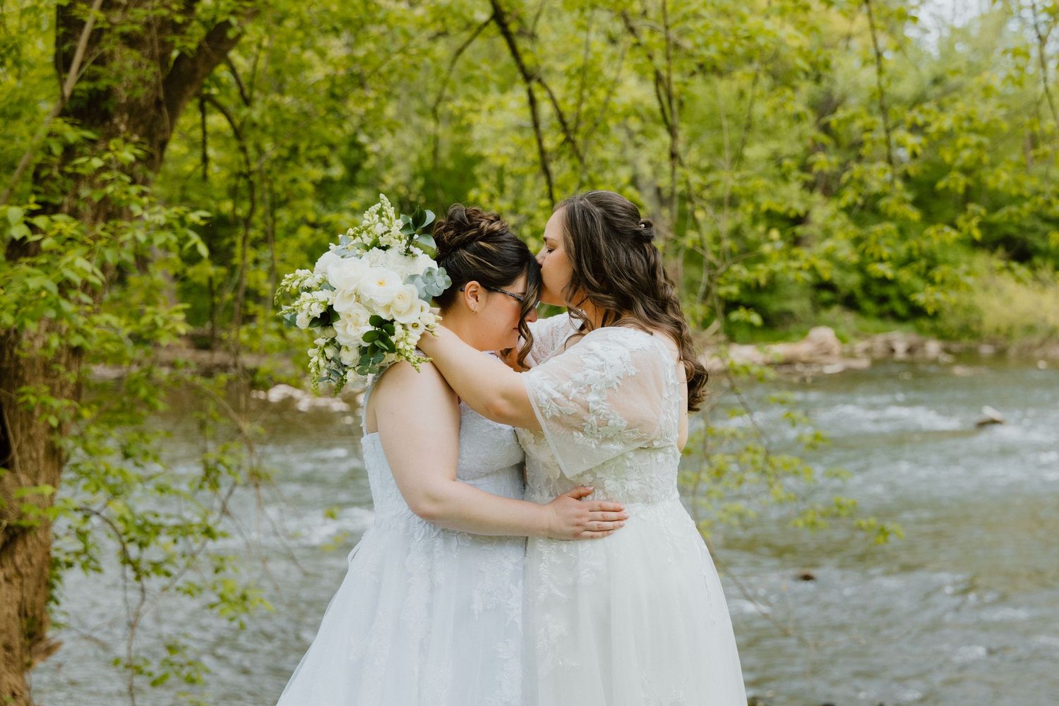 Two brides embracing by a wooded creek during an outdoor wedding.