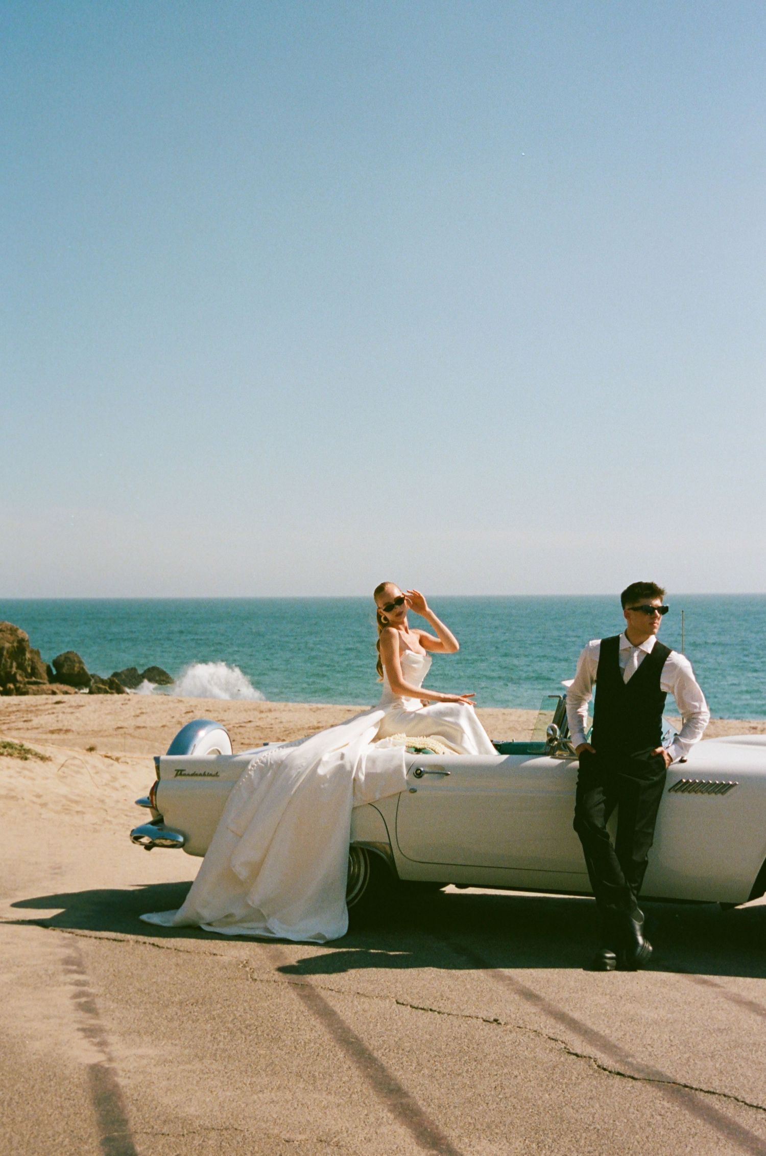 Bride and groom beside a vintage convertible on a coastal beach, captured in soft cinematic film tones.
