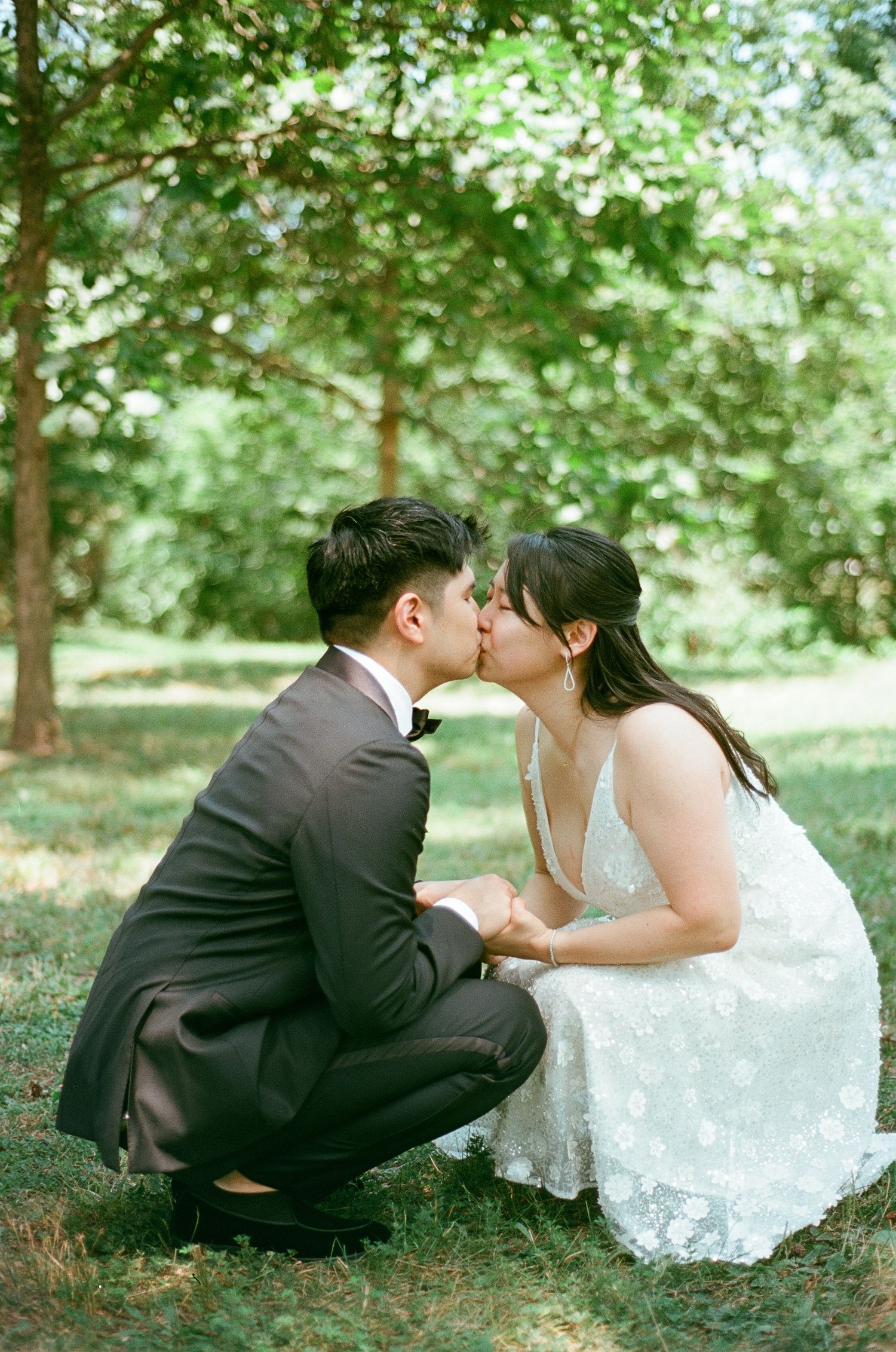 Bride and groom kissing while crouched together in a sunlit green park.