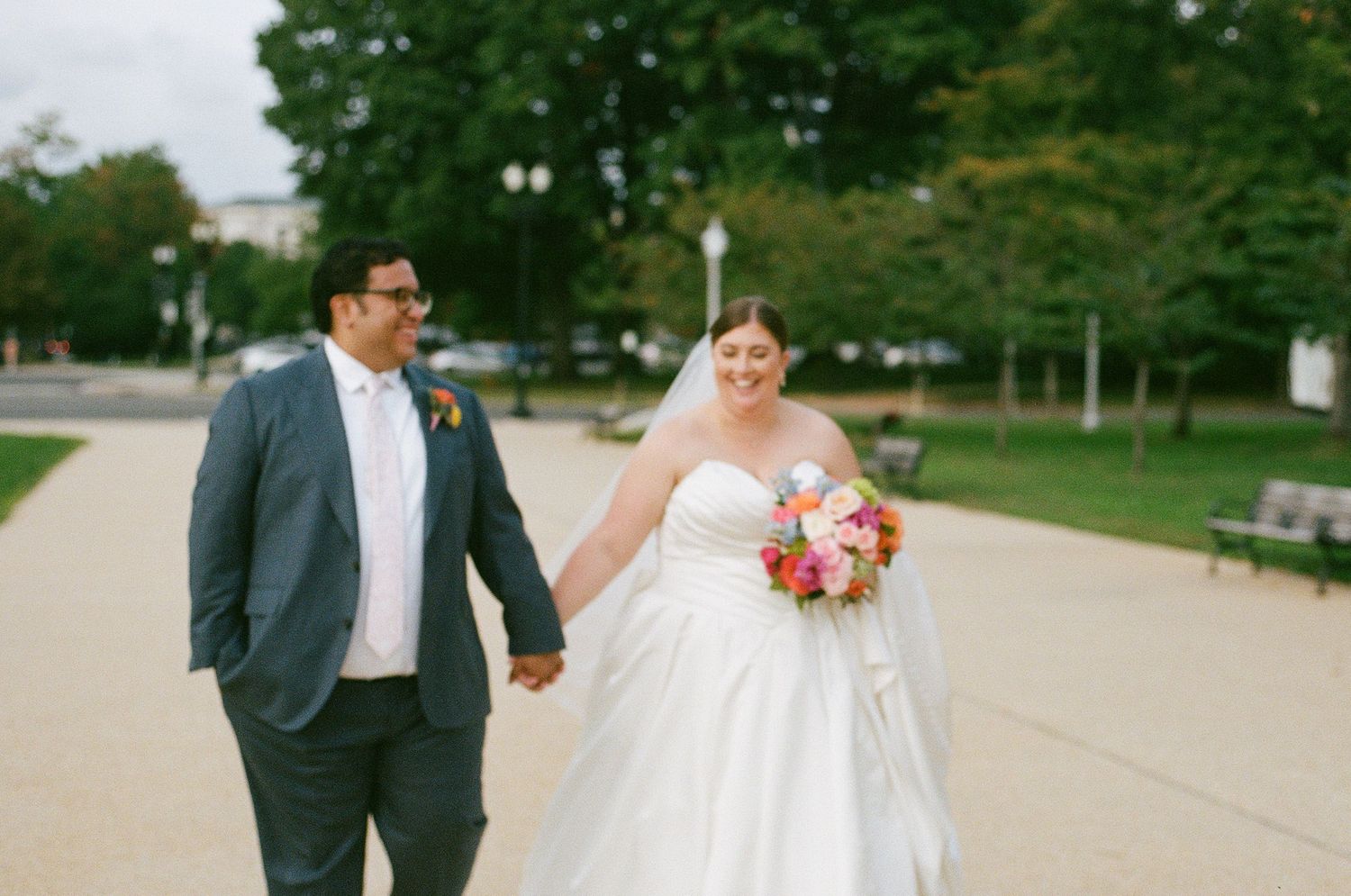 Bride and groom walking together along a brick pathway with a bouquet of red and orange blooms.