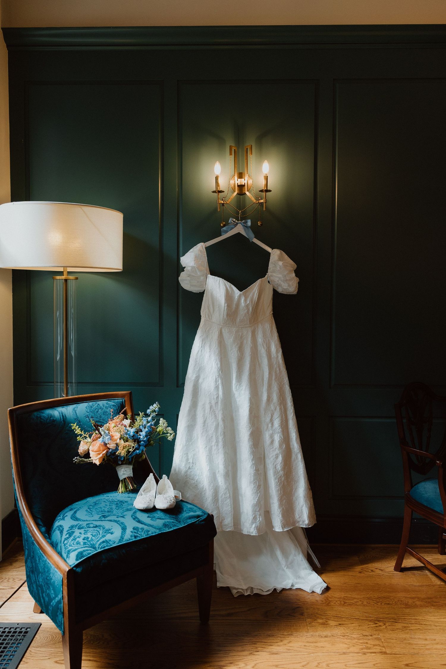 Bride's wedding gown hanging in a green-walled getting-ready room with a vintage lamp and blue armchair.
