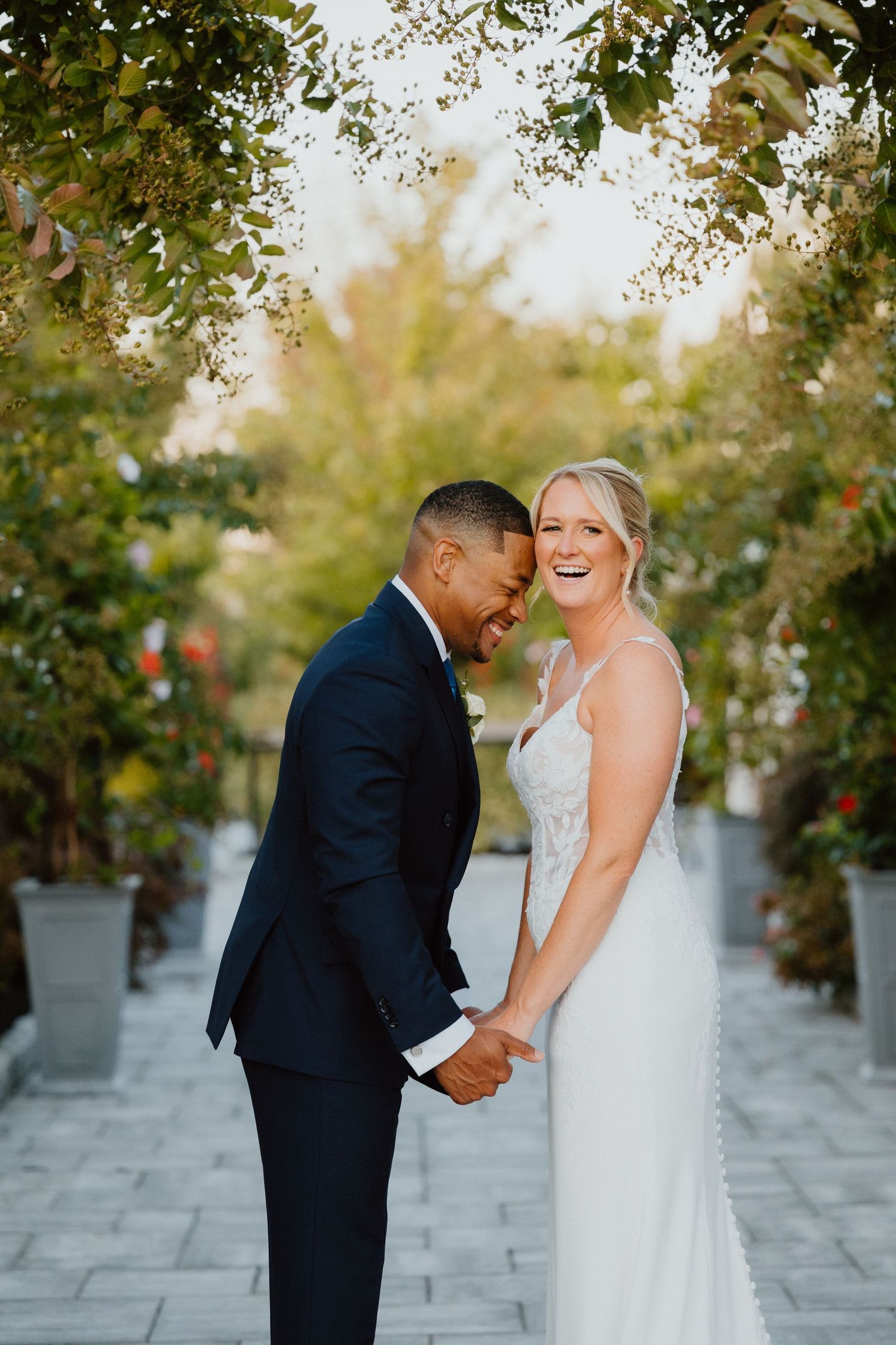 Bride and groom embracing forehead to forehead outdoors, framed by lush green trees and golden autumn light