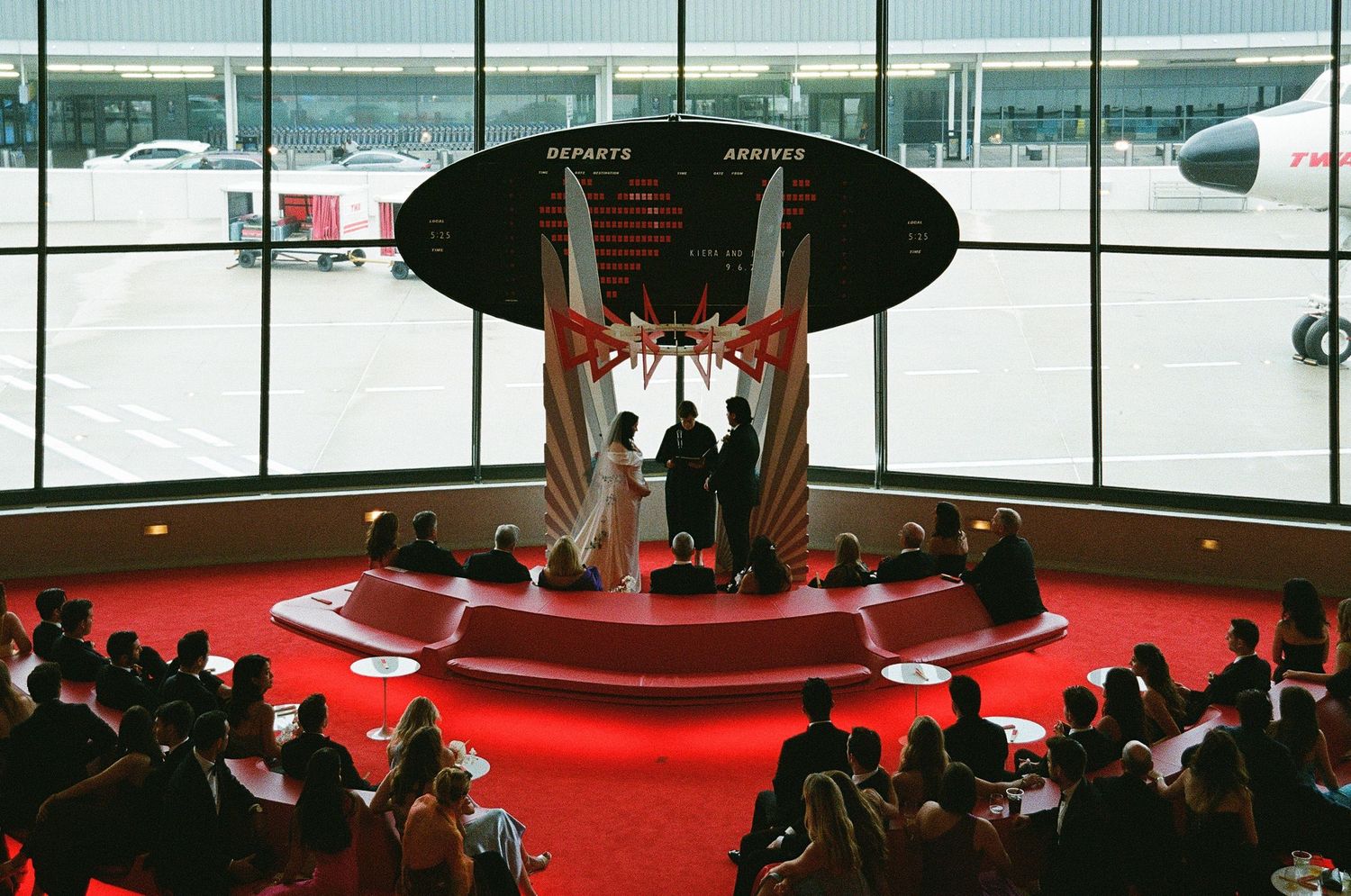 Wedding ceremony beneath the iconic curved mid-century architecture with guests gathered around a red-carpeted aisle.