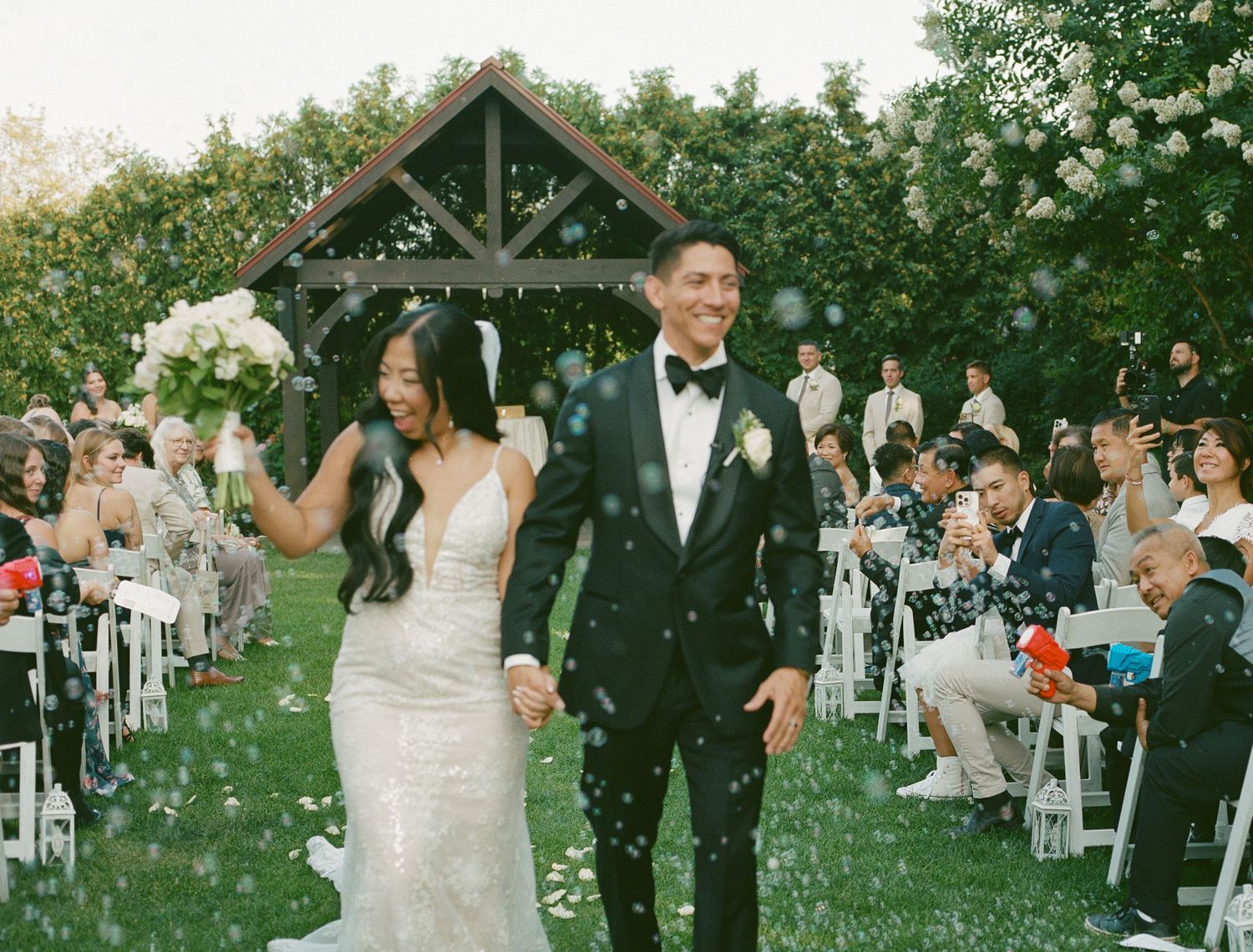 Bride and groom walking back down the aisle as guests cheer at an outdoor garden ceremony with a wooden arbor.