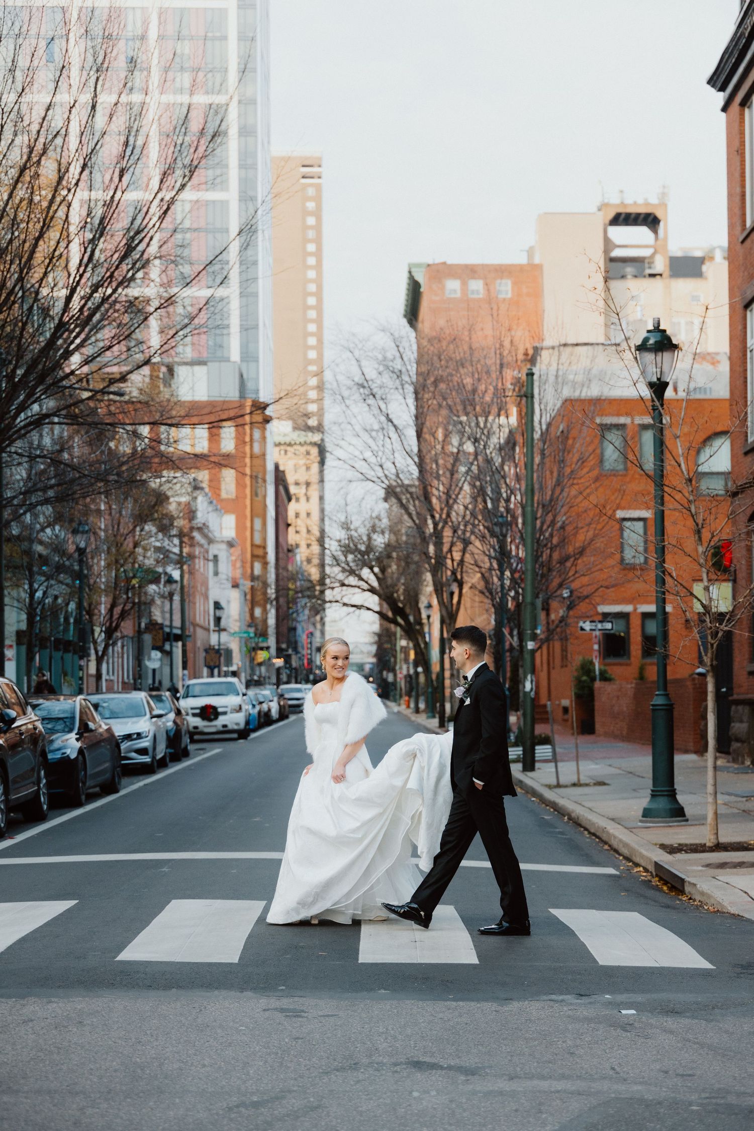 Bride walking across a city crosswalk between historic brick buildings on the wedding day.
