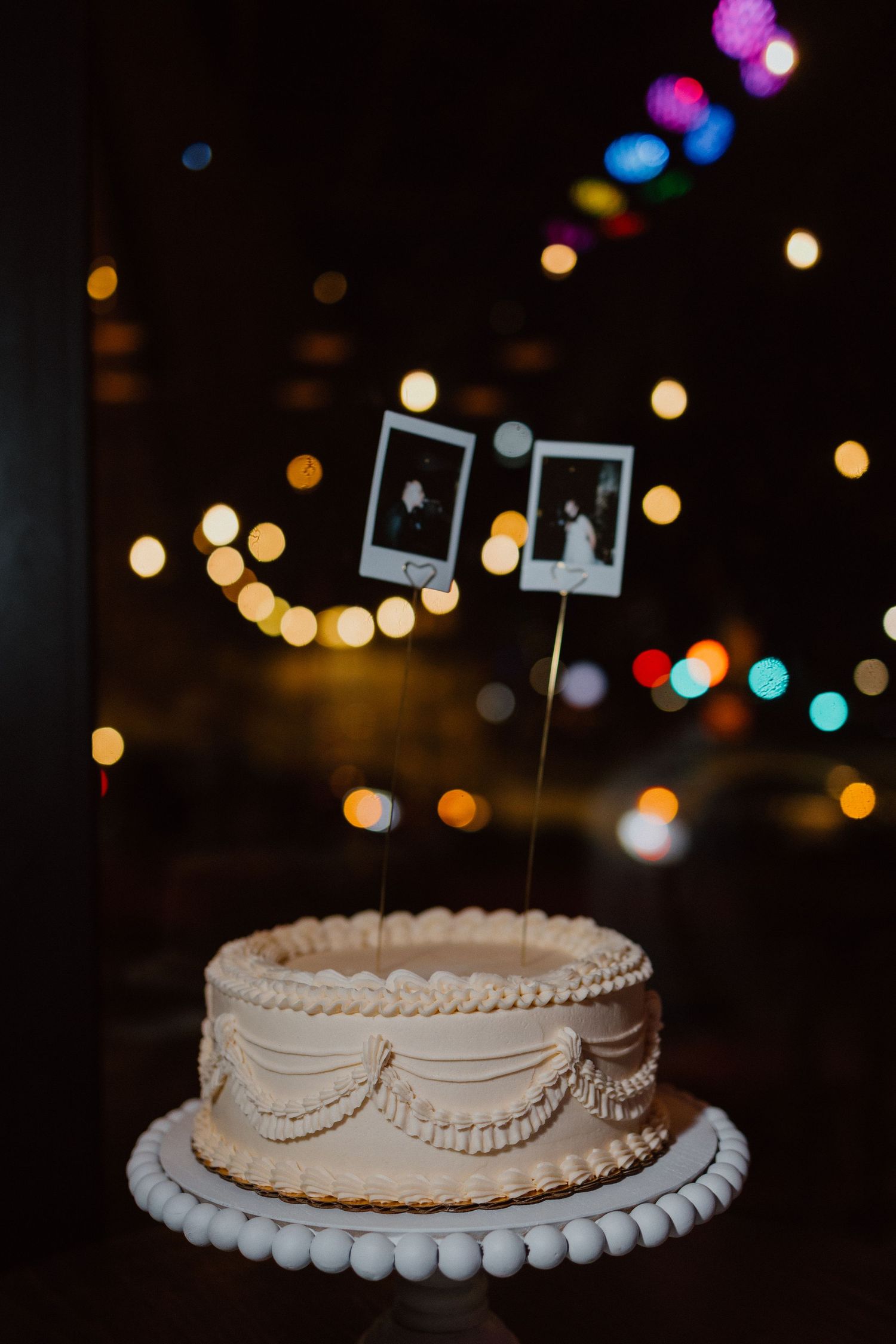 Small floral-topped wedding cake glowing softly under colorful twinkling reception lights.