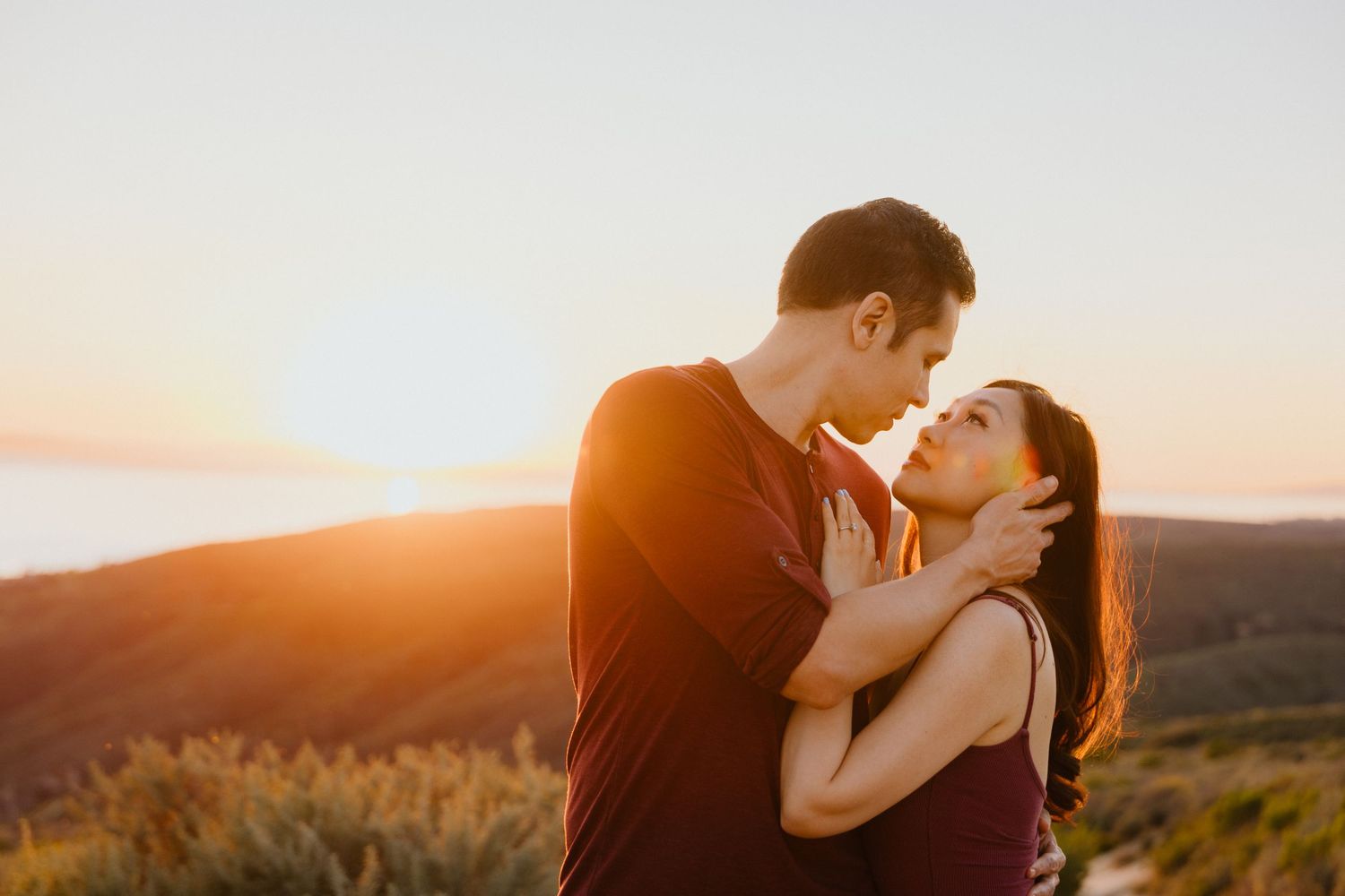 Couple sharing a tender embrace atop a hilltop bathed in warm sunset light, a romantic engagement moment.