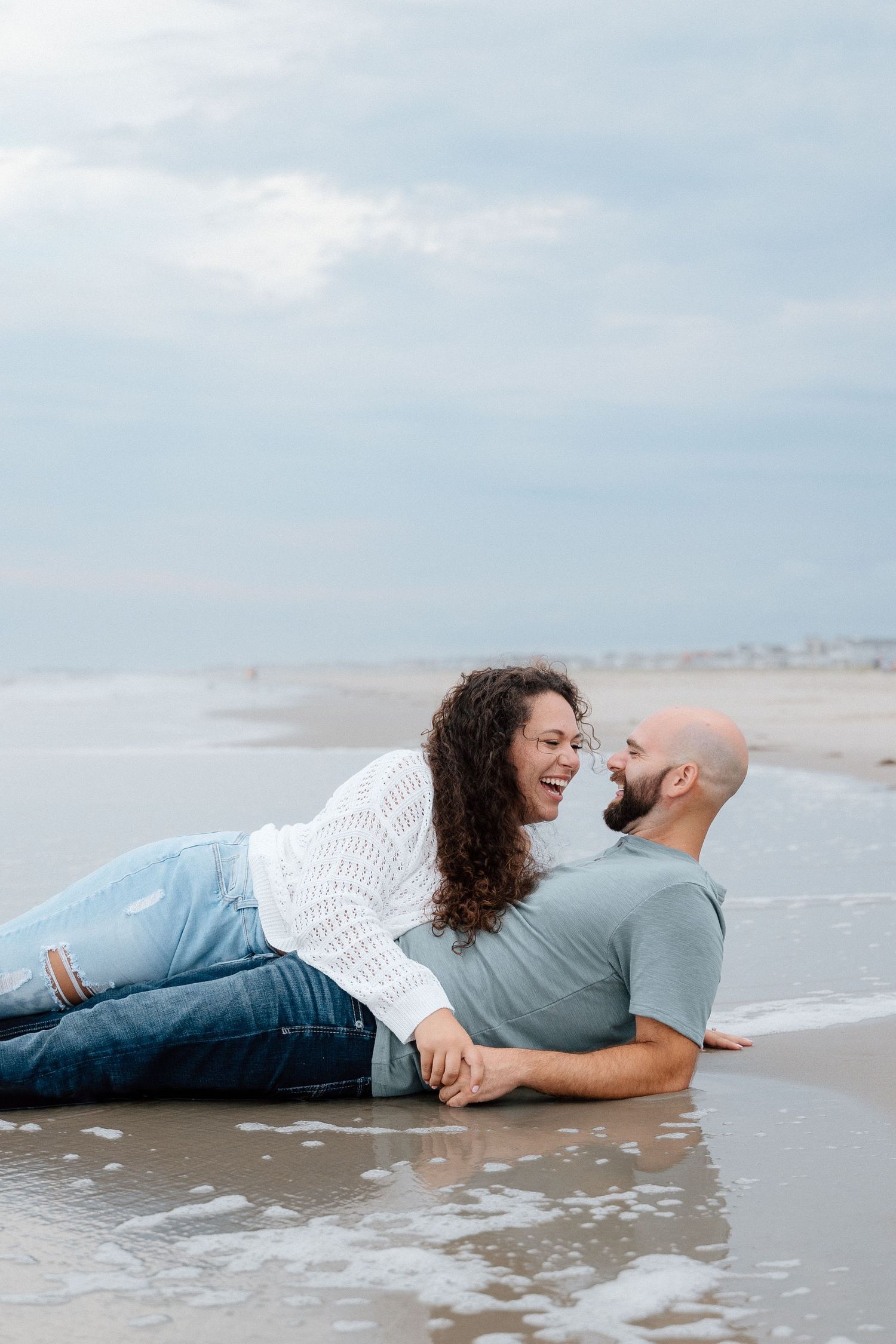 Couple lying together on the sand at the water's edge during a coastal engagement session.