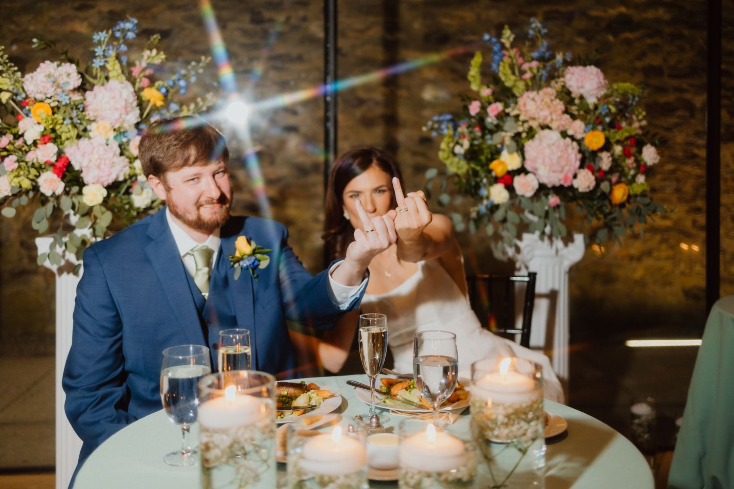 Newlyweds seated at a candlelit reception table beneath lush floral centerpieces.