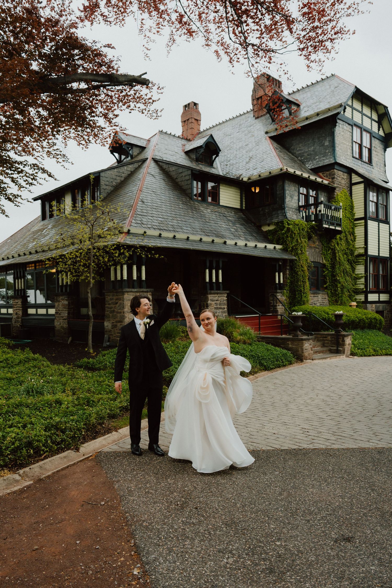 Bride and groom standing before a grand estate framed by autumn foliage.