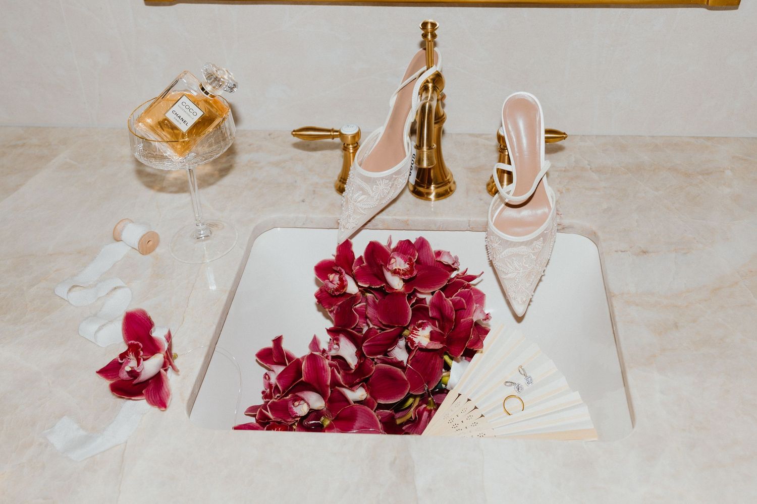 Wedding day flat lay with gold rings, perfume, and scattered red rose petals on marble.