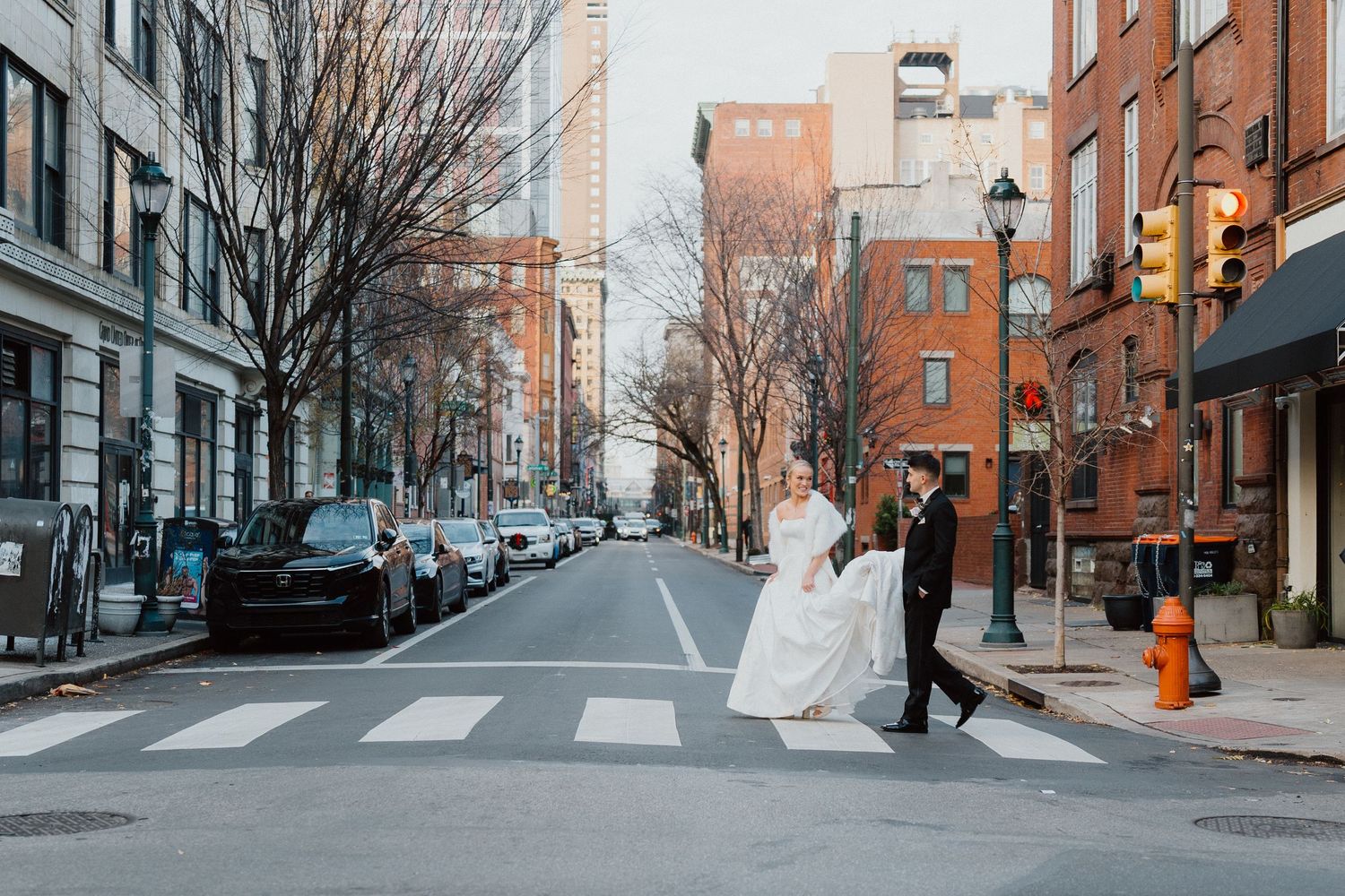 Bride and groom crossing a downtown crosswalk surrounded by brick buildings and autumn-toned trees.