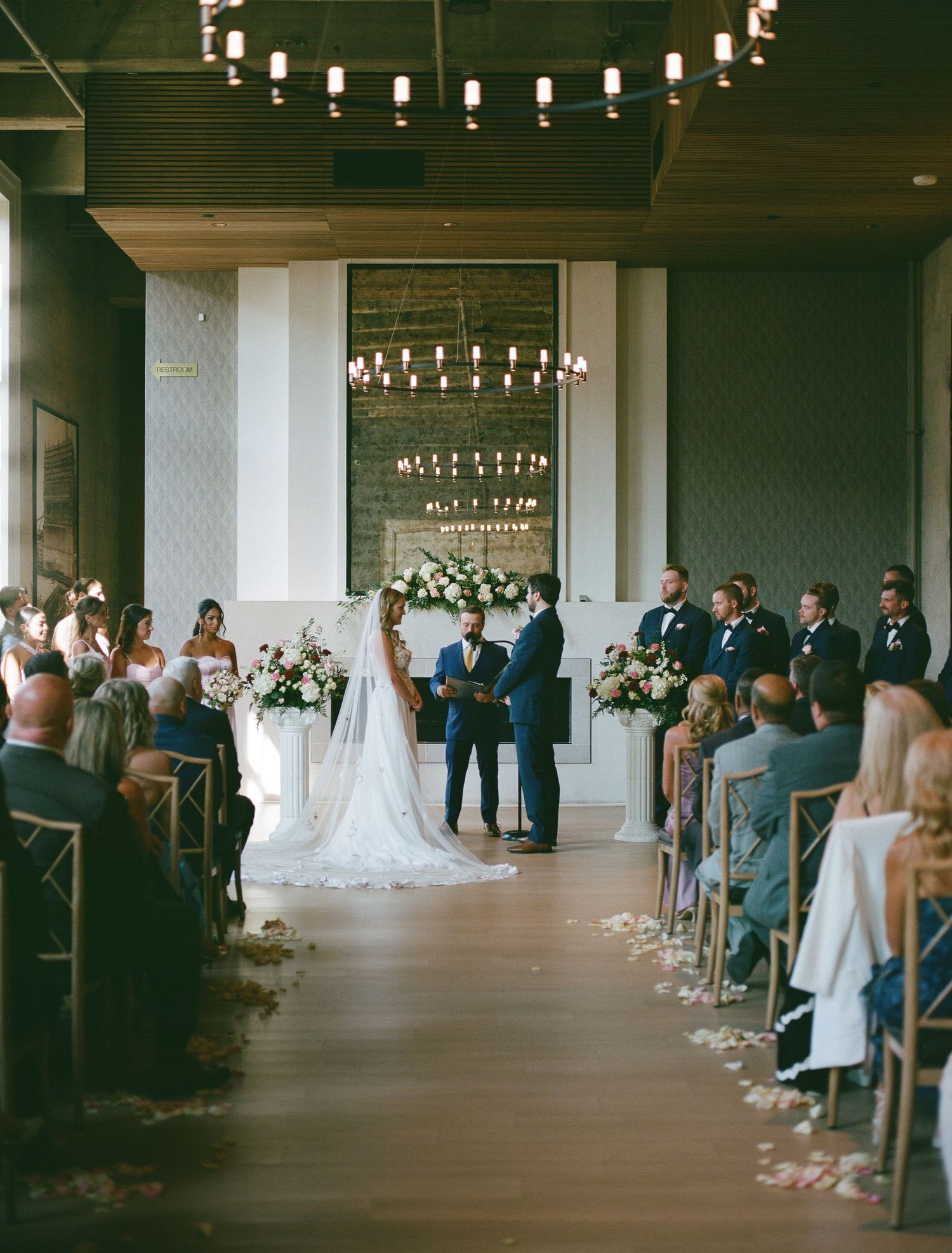 Wedding ceremony aisle view of guests gathered beneath string lights in an intimate covered venue.