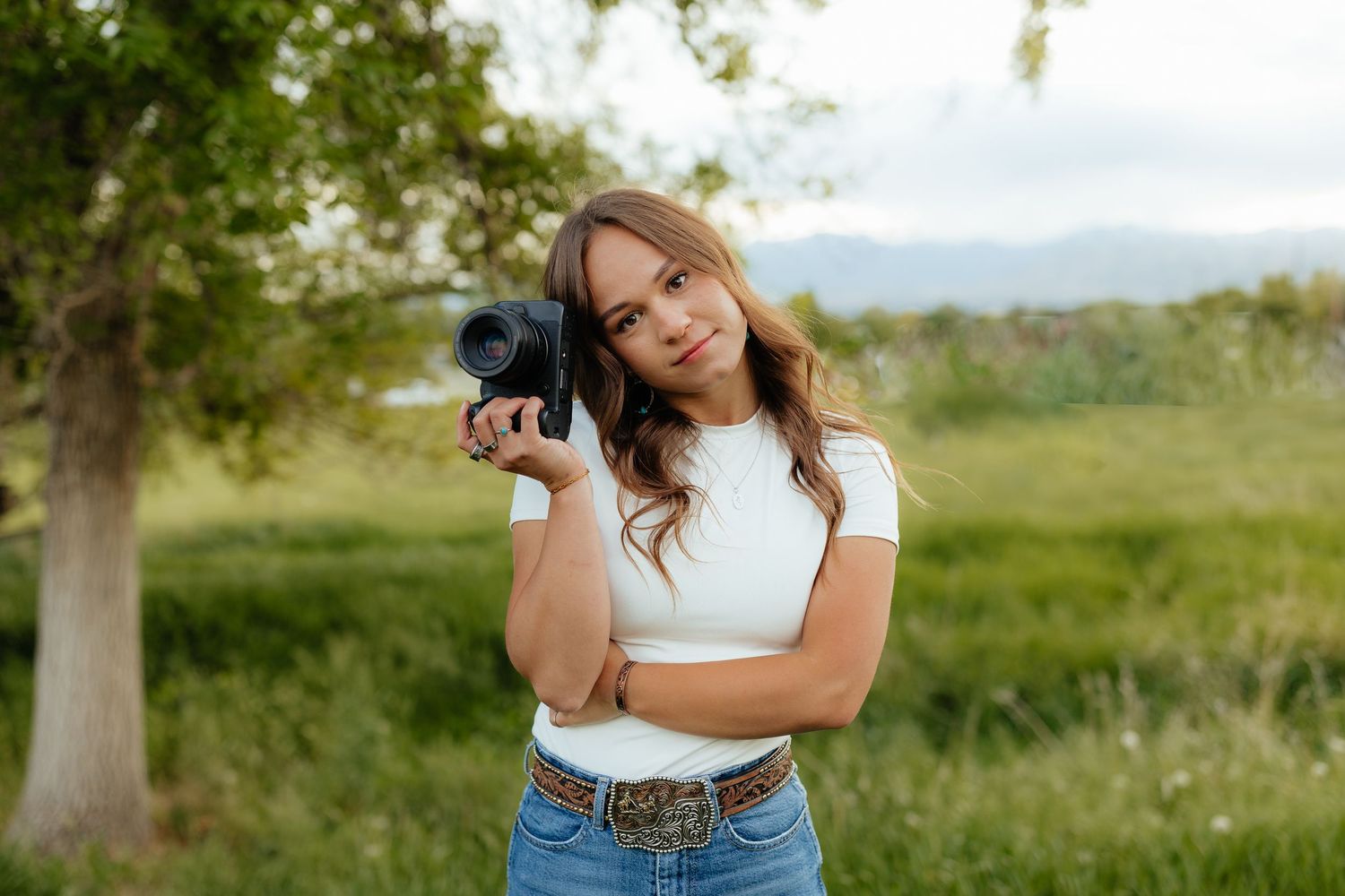 a western photographer holding up a camera to show she is the one taking pictures she is wearing a white shirt jeans and a belt buckle.
