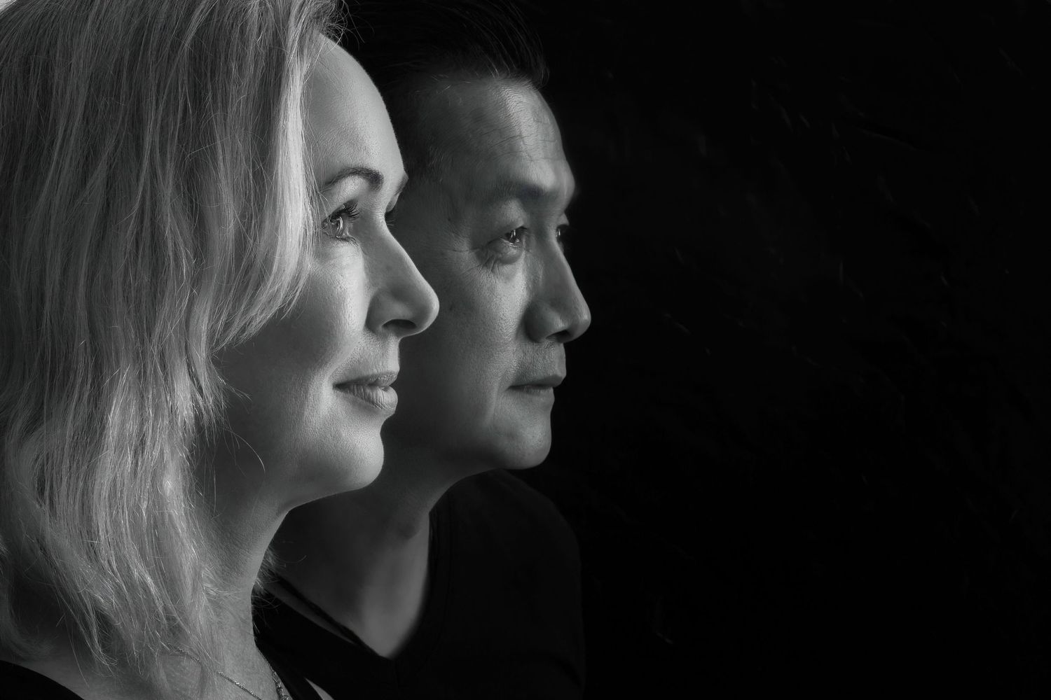 Black and white profile portrait of two people standing side by side with stark side lighting creating dramatic contrast.