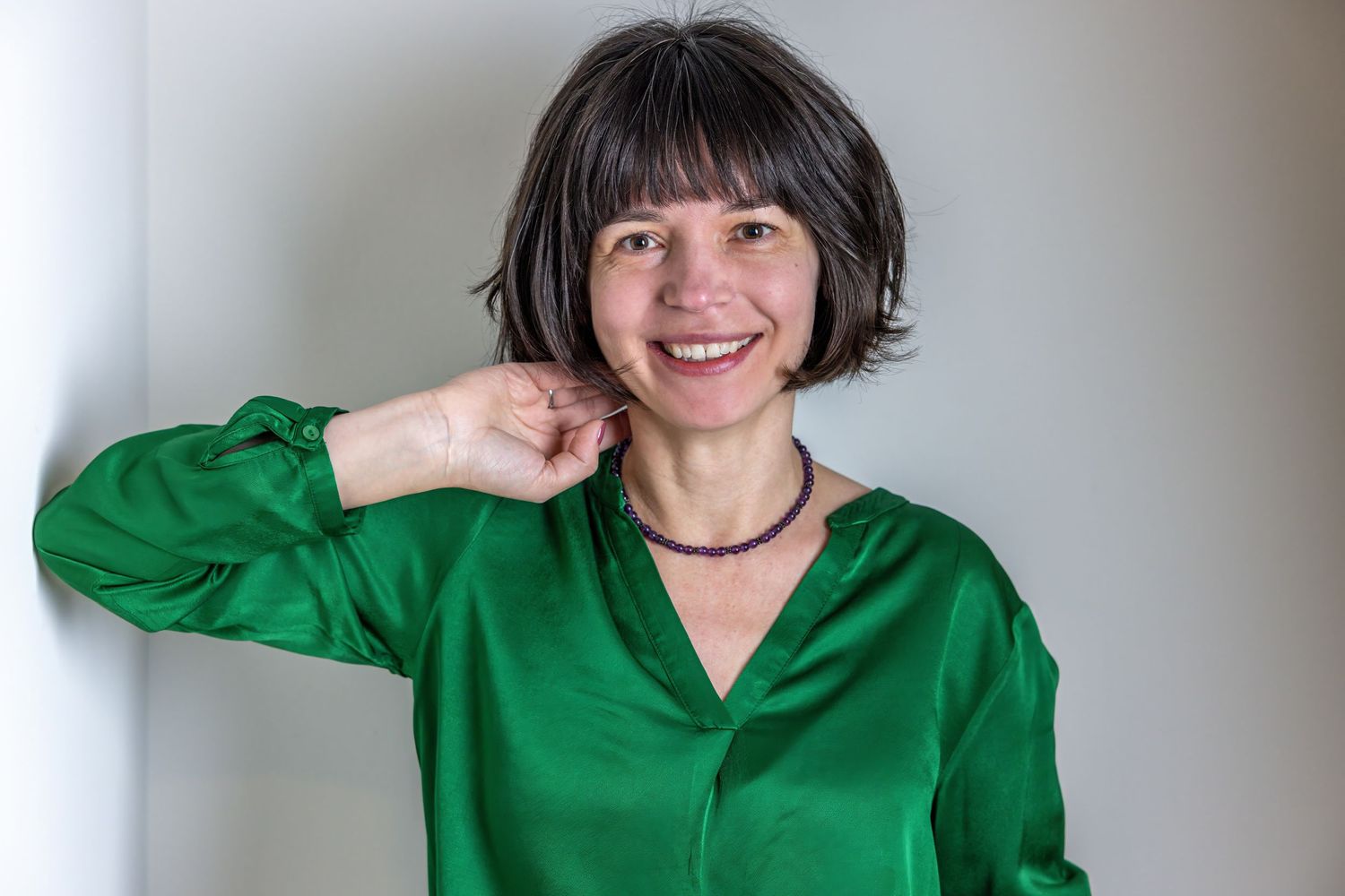 A person wearing a bright emerald green blouse smiles while touching their shoulder against a white background.