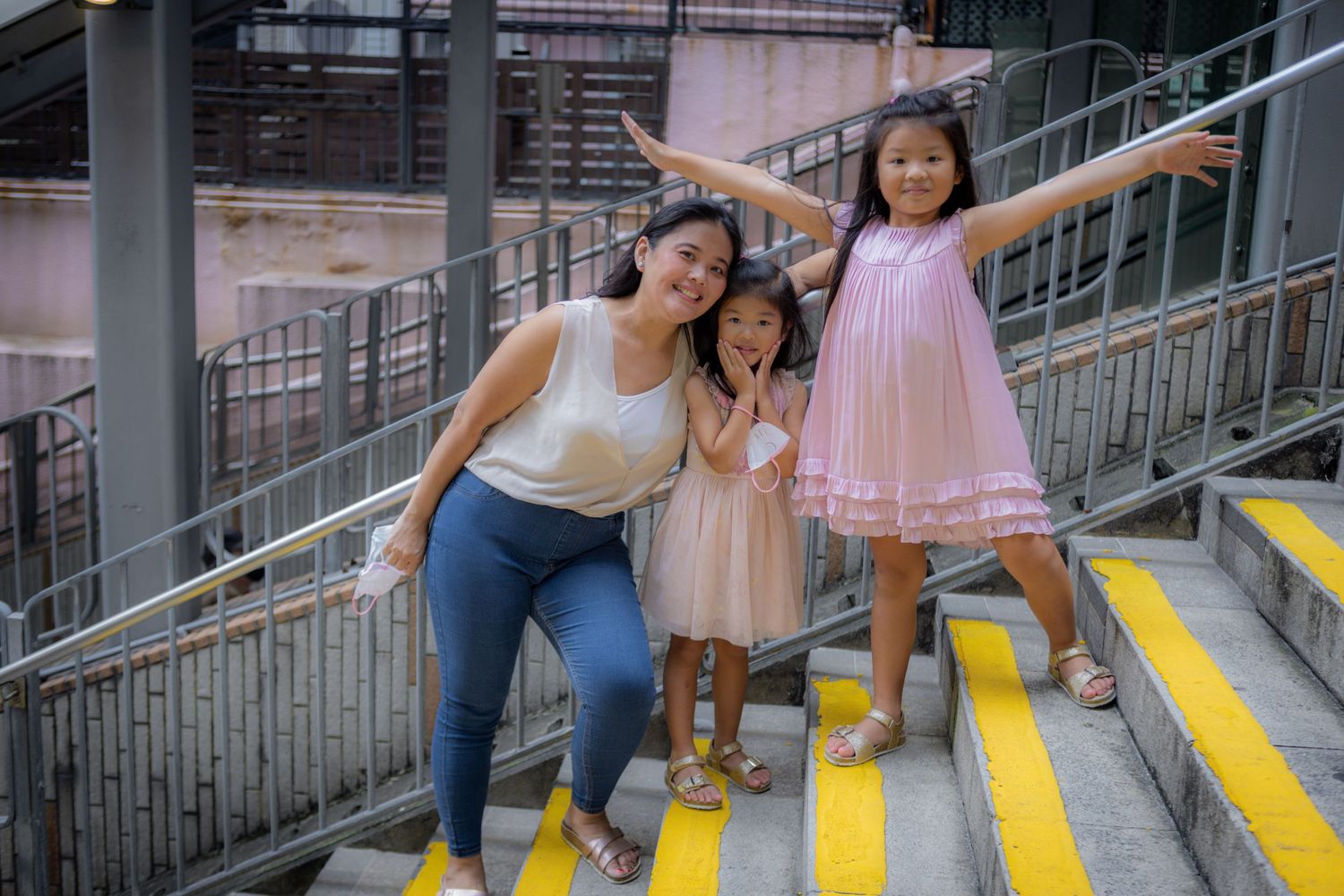 Mom and two young daughters stand joyfully with arms spread out on yellow-striped outdoor steps.