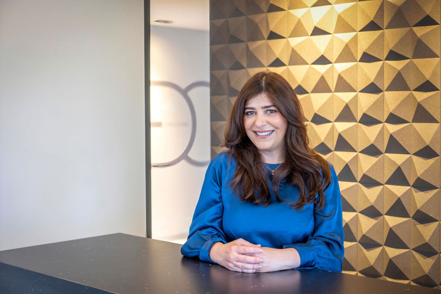 Professional photo showing someone in a bright blue sweater seated at a desk with a geometric gold wall pattern behind.