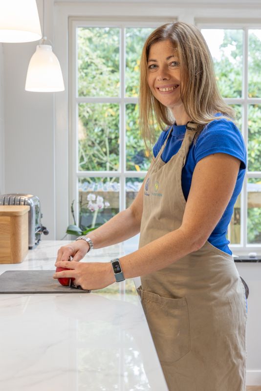 Wearing a blue shirt and beige apron, someone prepares food at a white kitchen counter near a bright window.