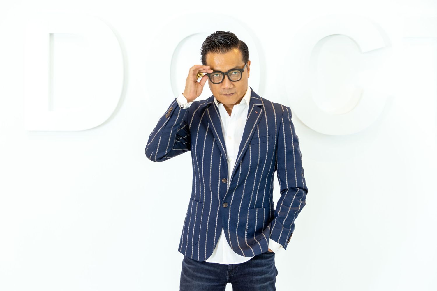 A professional Chinese man in a pinstriped navy business suit and glasses stands against a white background while adjusting their eyewear.