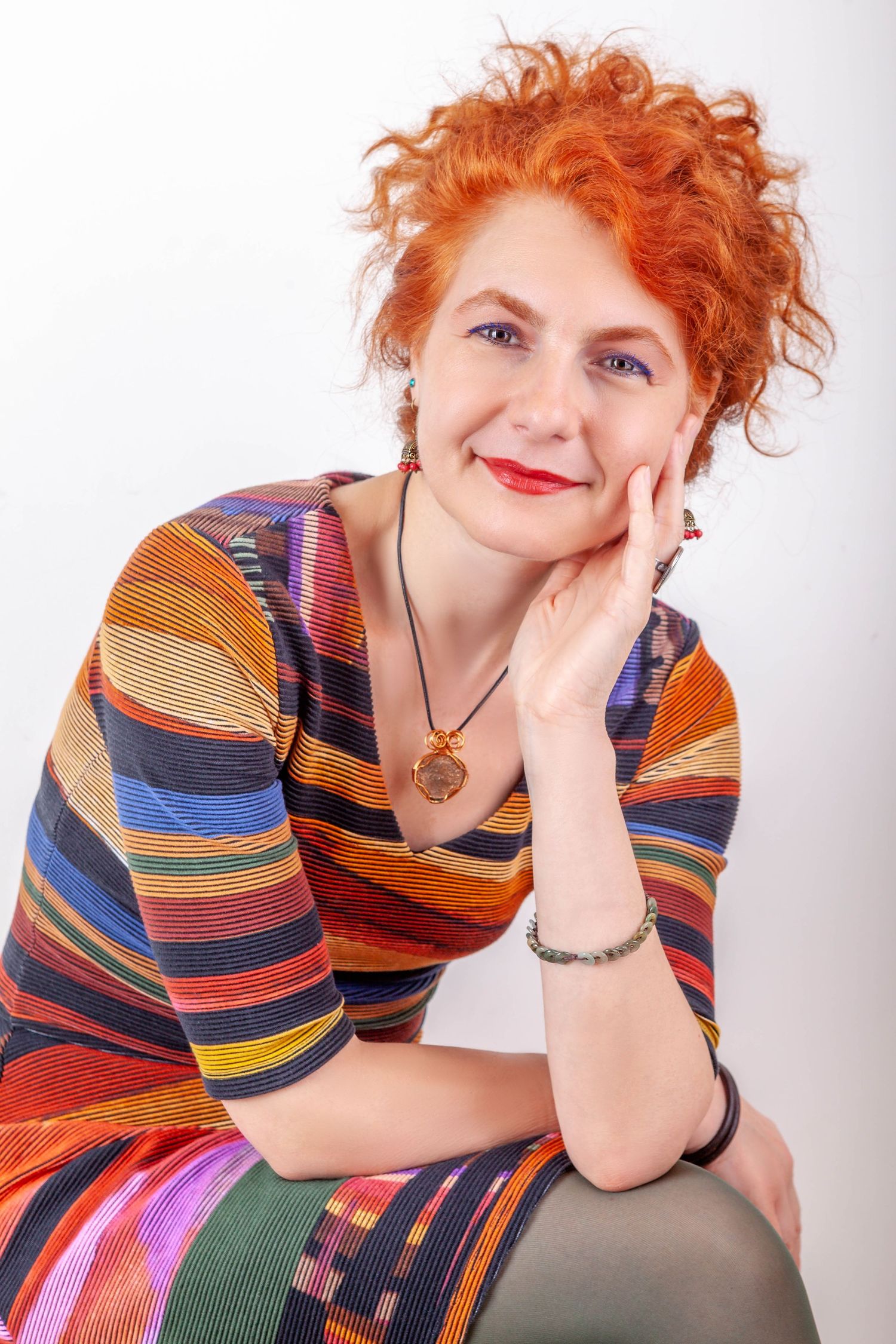 A person wearing a colorful striped dress with vibrant red curly hair poses against a white background.