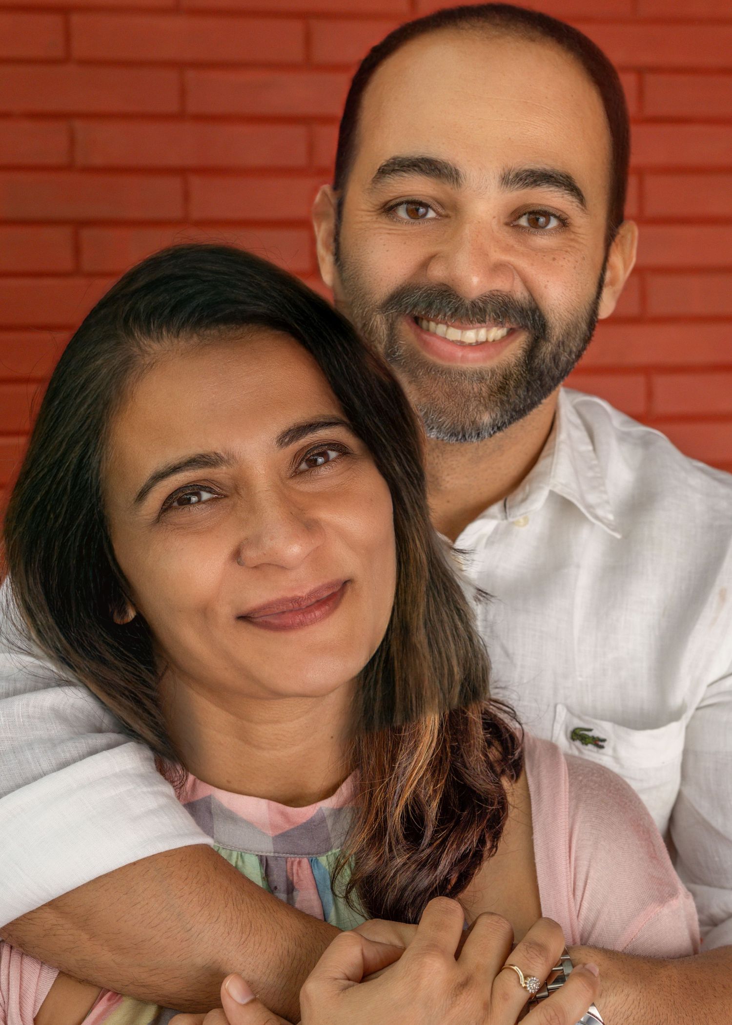 A happy couple in white and pink outfits smiling warmly against a rustic red brick wall background.