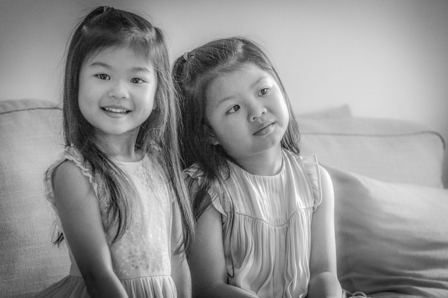 Two young girls in white sleeveless dresses sit together on a couch in a black and white portrait.