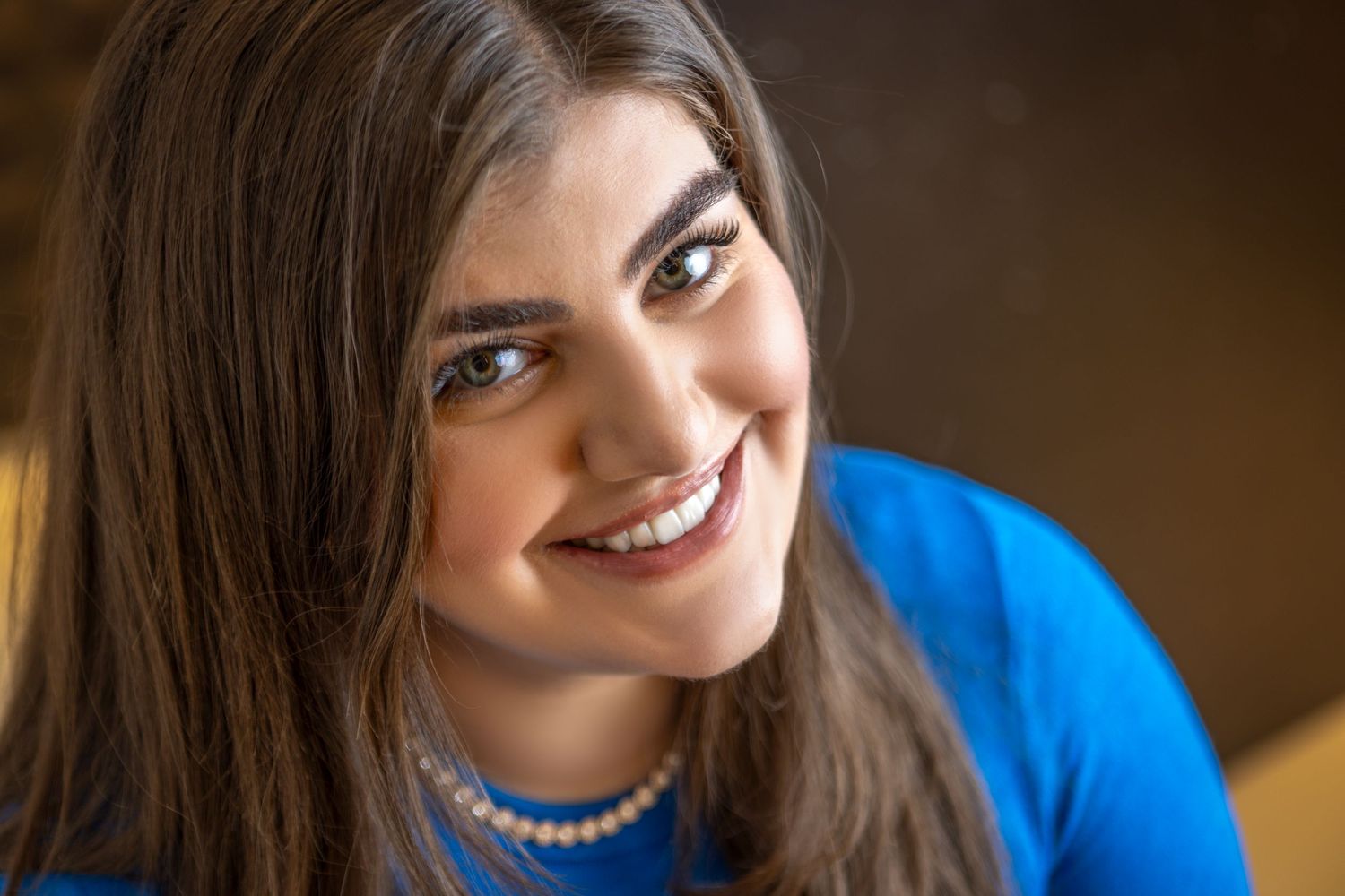 A bright smile stands out against a blue sweater and pearl necklace in a warm-toned portrait.