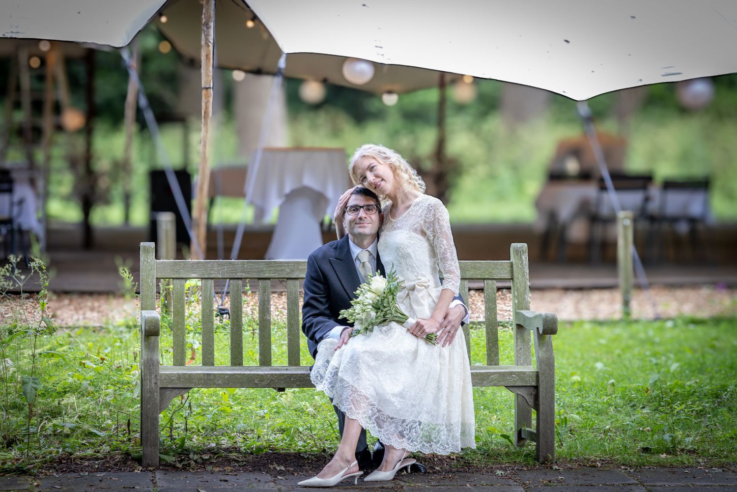 Two people share a tender moment on a wooden bench in a garden setting while holding a bouquet of flowers.